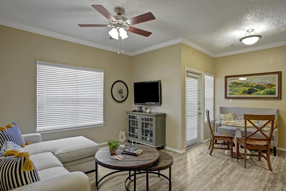 Arbors at Wolf Pen Creek Apartments Cozy living room with a sectional sofa, round tables, ceiling fan, dining area, and wall-mounted TV.