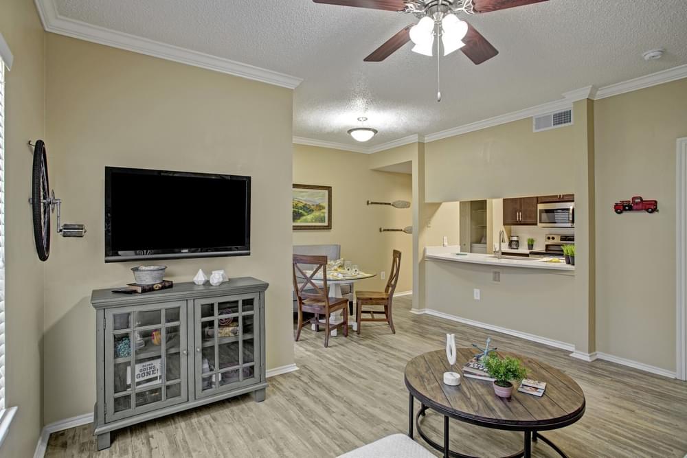 Arbors at Wolf Pen Creek Apartments Cozy living room with a sectional sofa, round tables, ceiling fan, dining area, and wall-mounted TV.