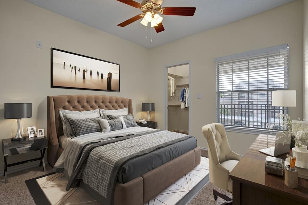 Arbors at Wolf Pen Creek Apartments bedroom with large bed, wooden desk, upholstered office chair, large window, closet, and ceiling fan.