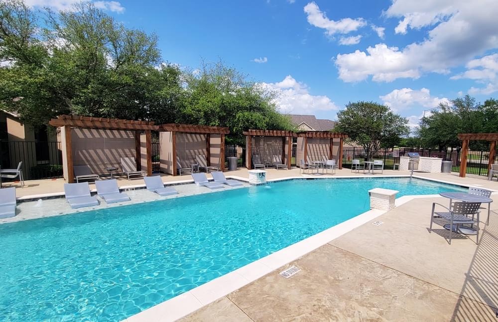 Arbors at Wolf Pen Creek Apartments Outdoor swimming pool with loungers and shaded pergolas on a sunny day, surrounded by trees and a clear blue sky.
