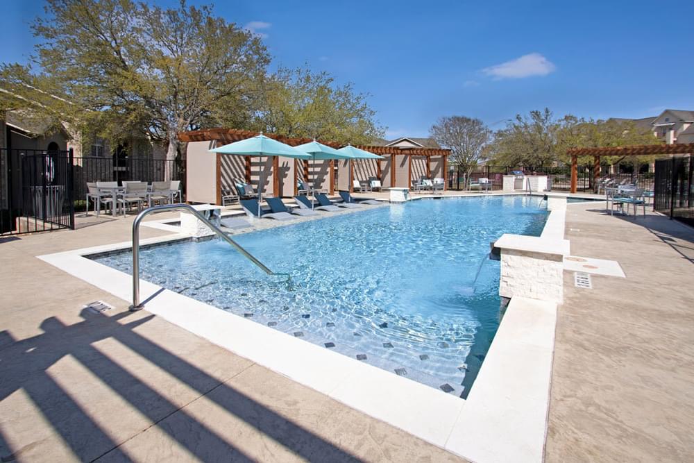 Arbors at Wolf Pen Creek Apartments Outdoor swimming pool with loungers and shaded pergolas on a sunny day, surrounded by trees and a clear blue sky.