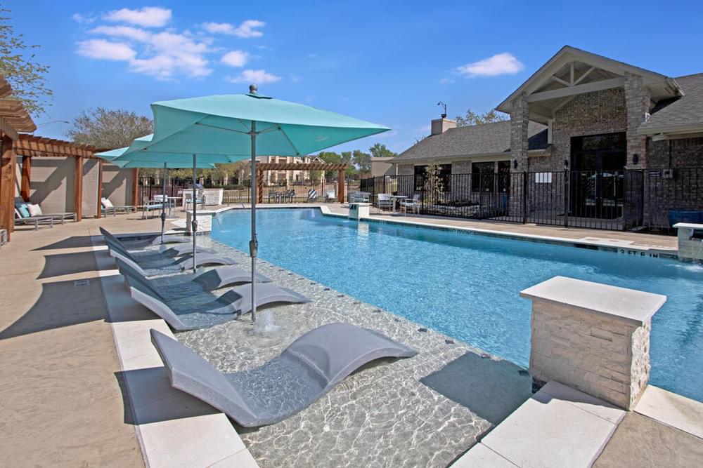 Arbors at Wolf Pen Creek Apartments Outdoor swimming pool with loungers and shaded pergolas on a sunny day, surrounded by trees and a clear blue sky.