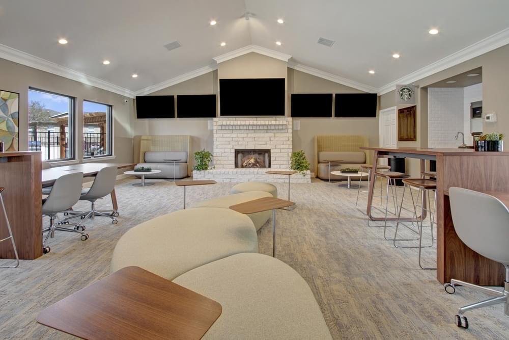 Arbors at Wolf Pen Creek Apartments modern lounge with cozy seating, a fireplace, and large TVs on the wall under a vaulted ceiling. Neutral tones.