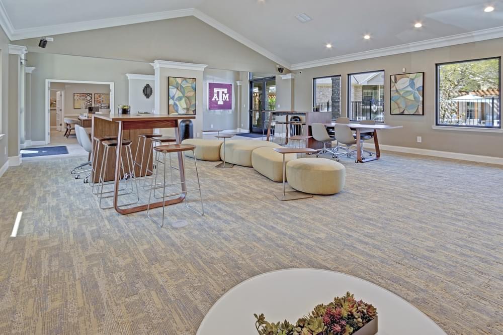 Arbors at Wolf Pen Creek Apartments modern lounge with cozy seating, a fireplace, and large TVs on the wall under a vaulted ceiling. Neutral tones.