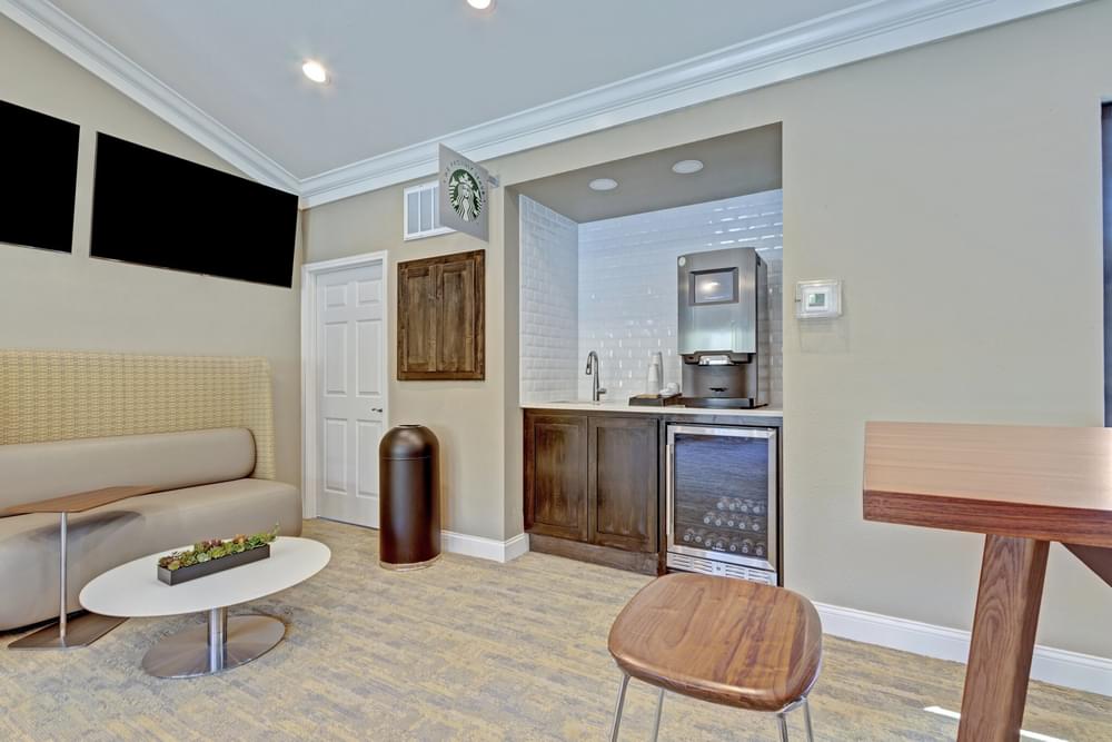 Arbors at Wolf Pen Creek Apartments modern lounge with cozy seating, a fireplace, and large TVs on the wall under a vaulted ceiling. Neutral tones.