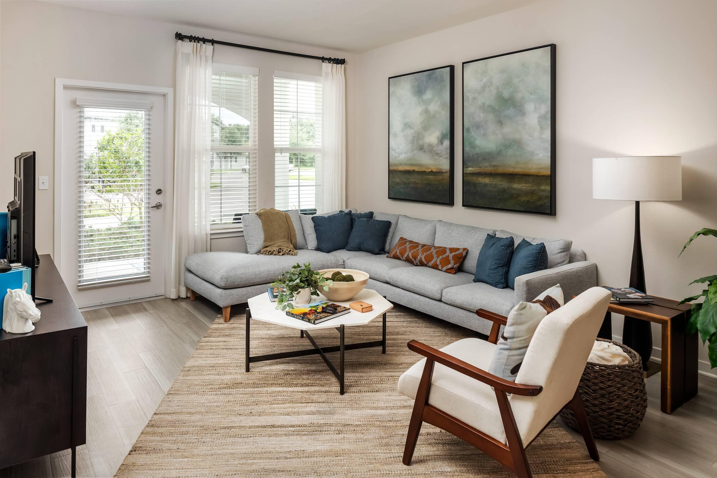 Shadetree Apartments living room with an L-shaped sofa, a chair, coffee table, rug, wall art, and large windows letting in natural light