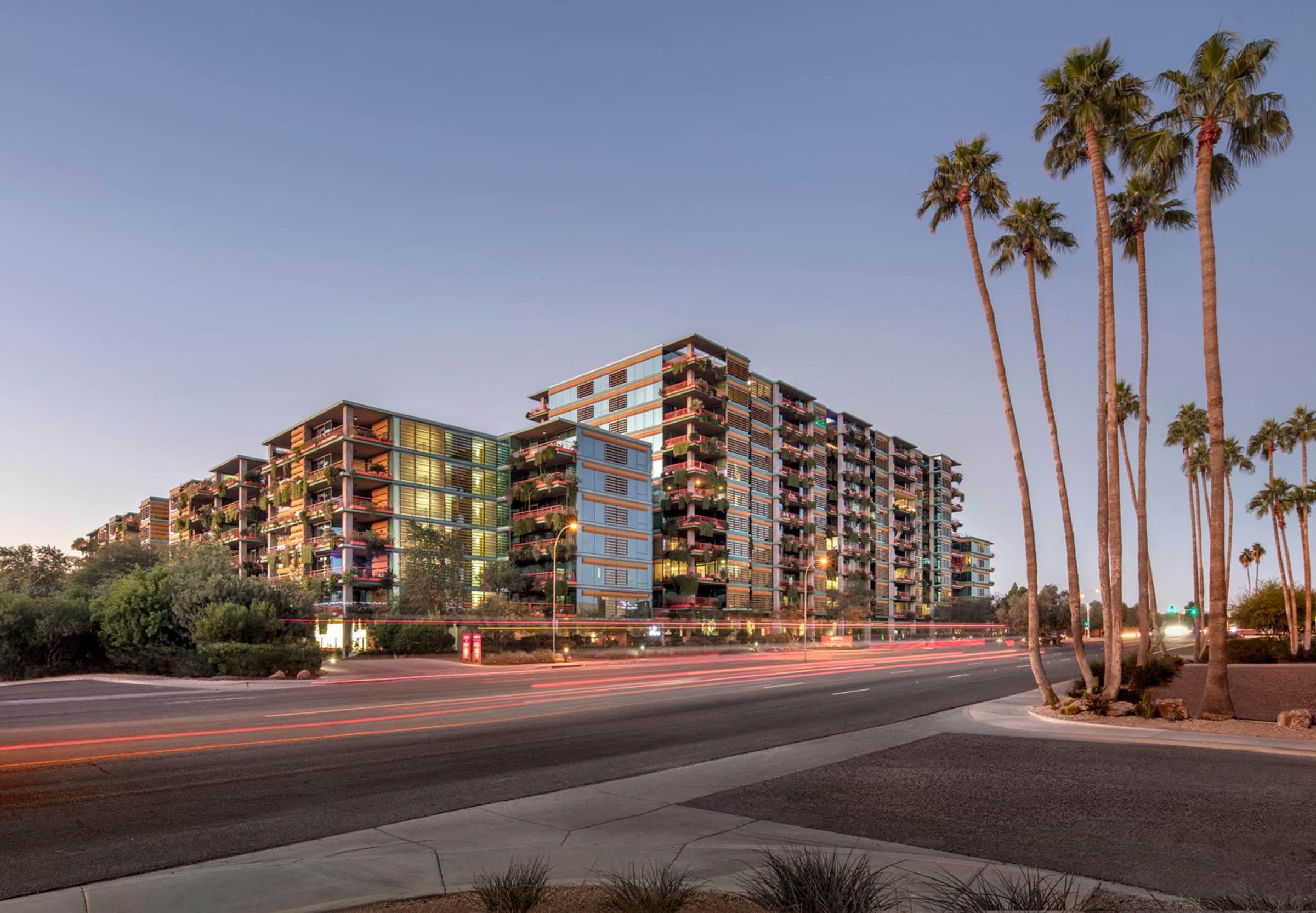 Optima Sonoran Village modern multi-story apartment building with balconies, against a dusk sky. Street and light trails in the foreground.