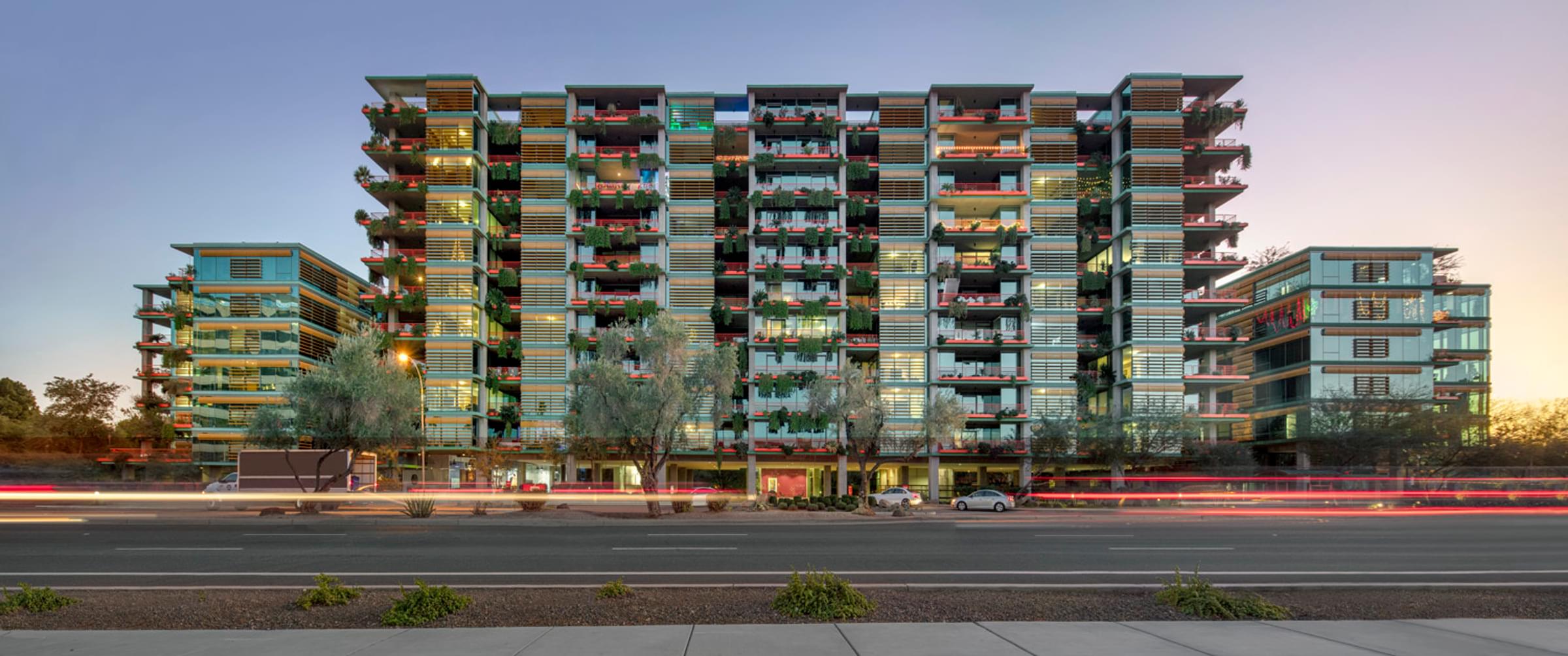 Optima Sonoran Village modern multi-story apartment building with balconies, against a dusk sky. Street and light trails in the foreground.