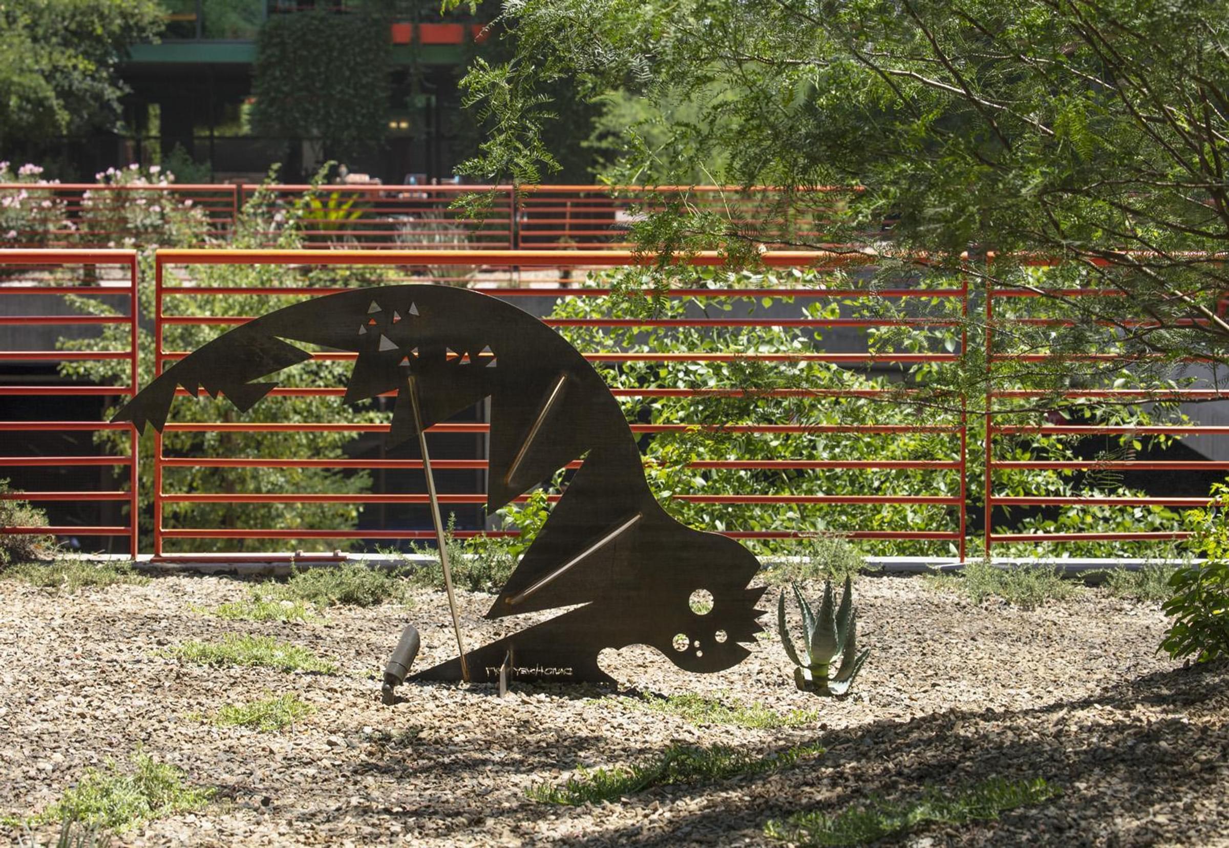 Optima Sonoran Village apartments Metal fish sculpture near red fence, surrounded by trees and plants in a landscaped outdoor area.