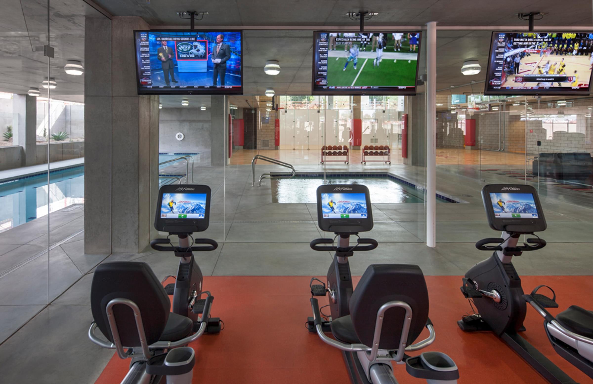 A modern gym with stationary bikes, wall-mounted TVs, and a pool view through glass panels at Optima Sonoran Village apartments.