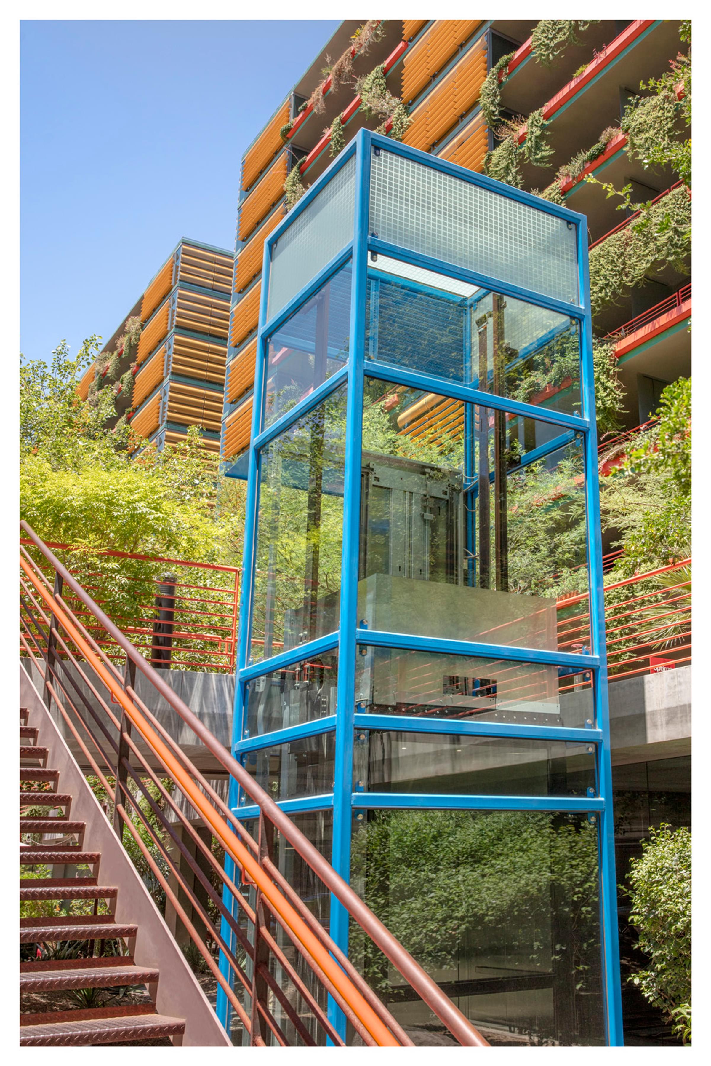 Optima Sonoran Village apartments Outdoor glass elevator with a blue frame next to stairs, surrounded by greenery and a colorful building facade.