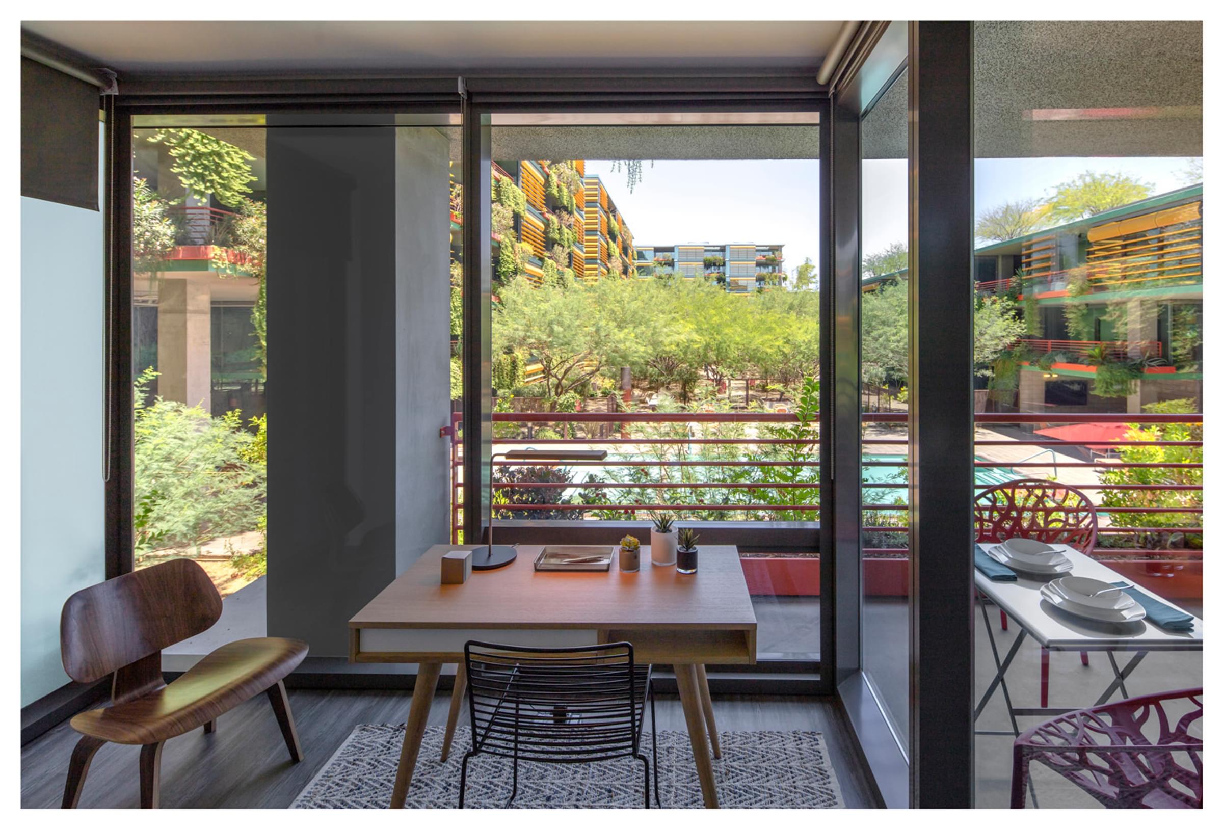 Optima Sonoran Village apartments Modern room with a desk and chair, overlooking a garden and colorful buildings through large glass windows.