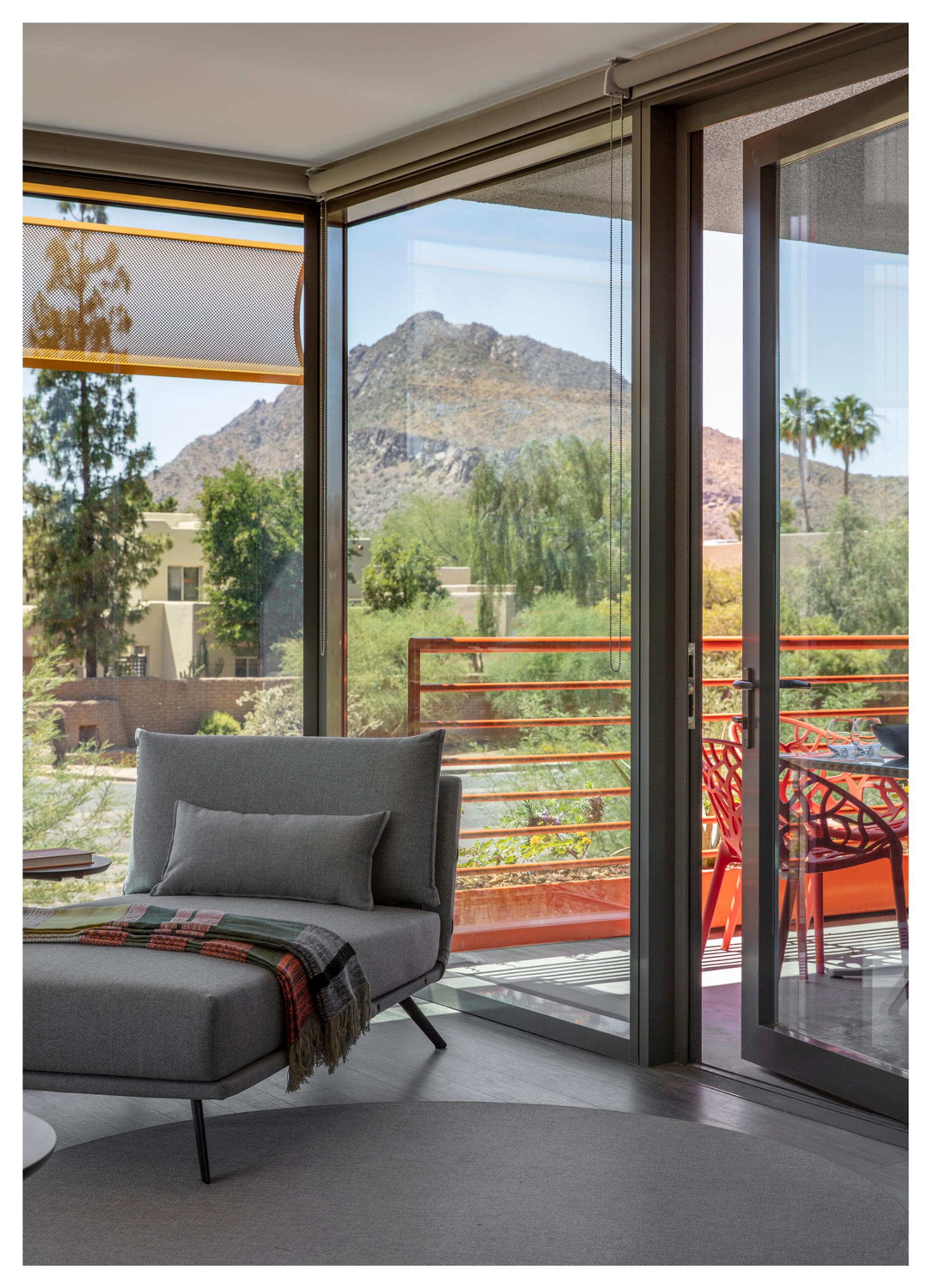 Optima Sonoran Village apartments Sunny room with gray chair, blanket, open glass doors leading to a balcony with red chairs, mountain view outside.