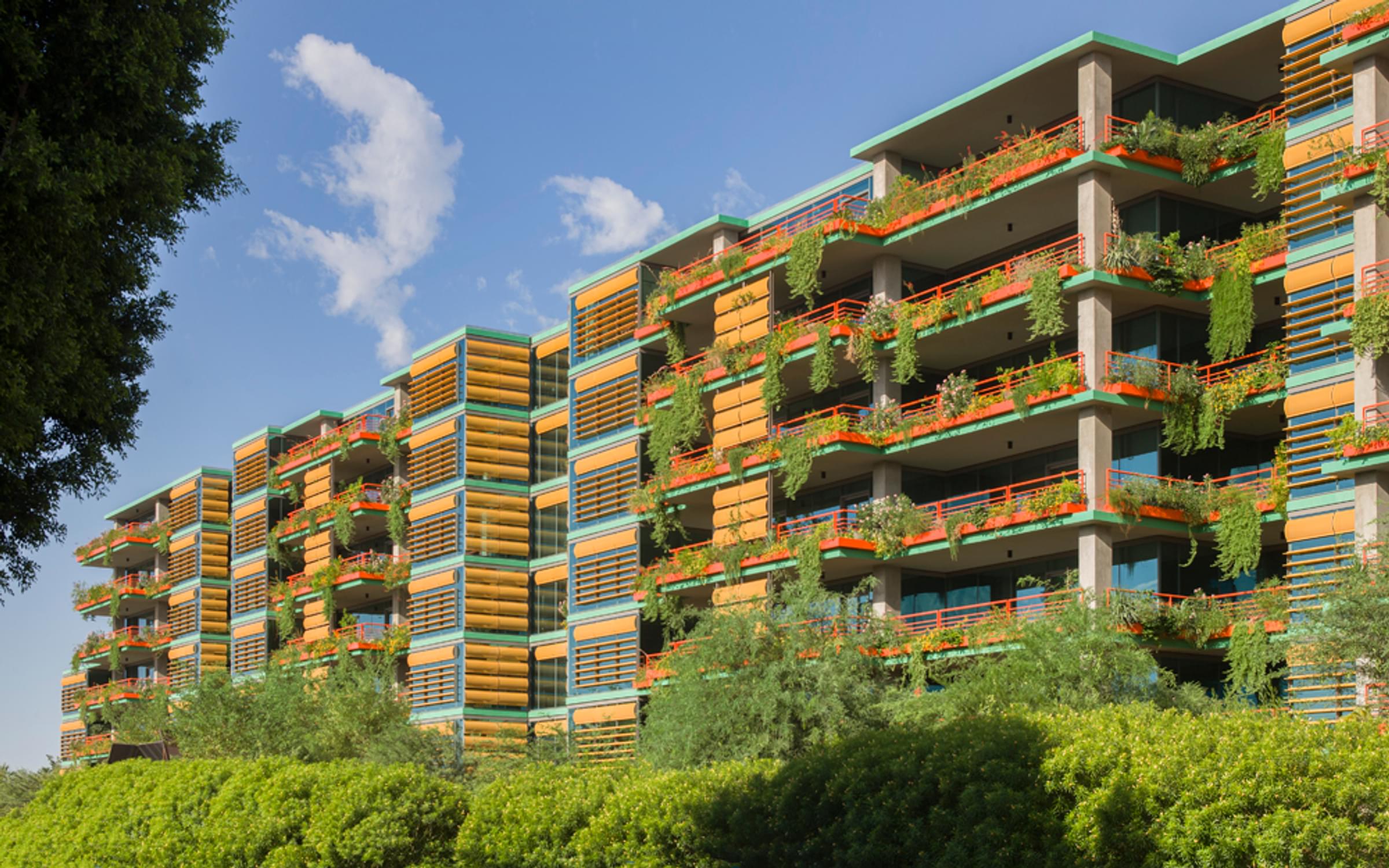 Optima Sonoran Village modern apartment building with colorful accents and lush plants on balconies, under a clear blue sky.
