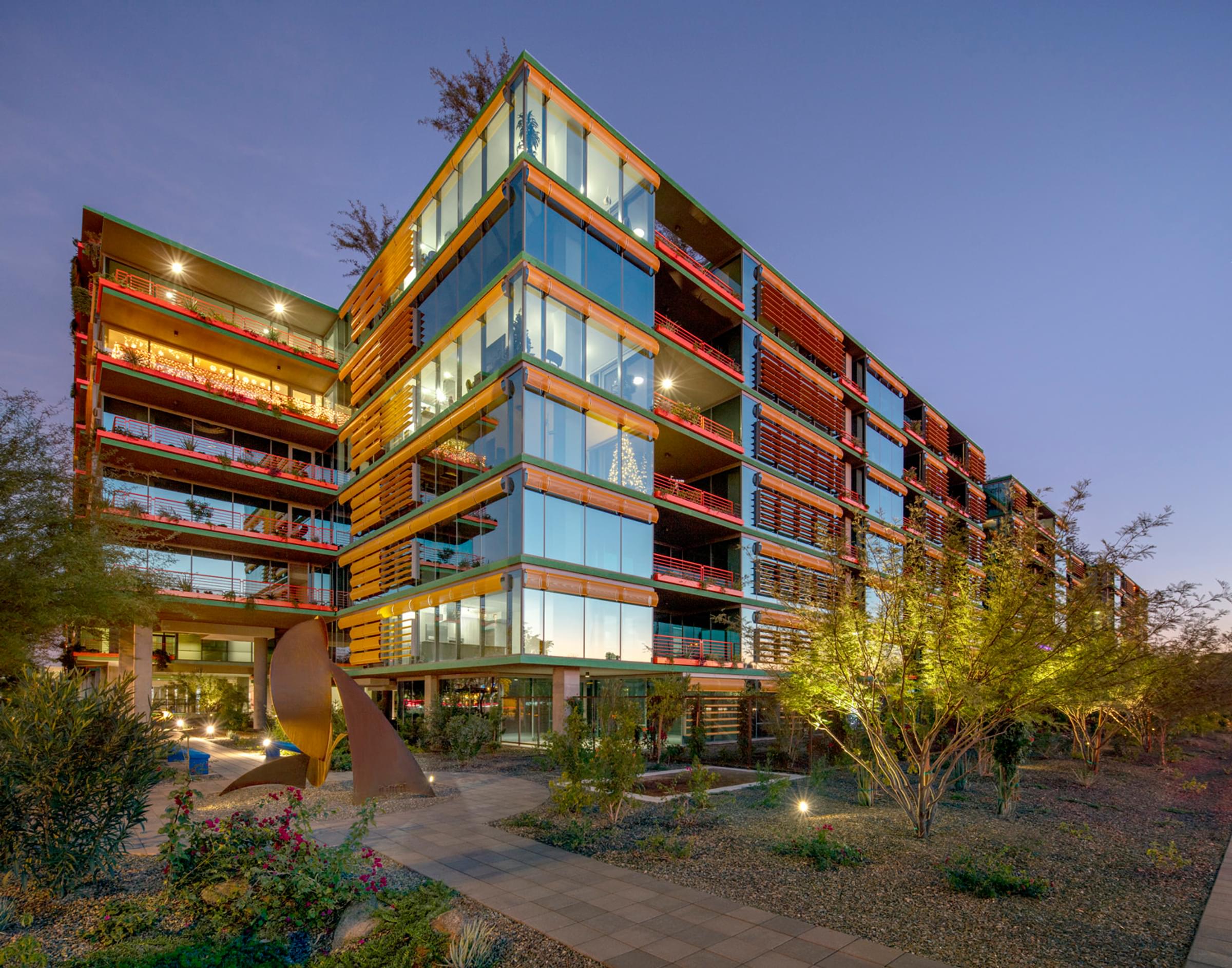 Optima Sonoran Village Modern glass apartment building surrounded by desert landscaping at dusk, with lights on inside.