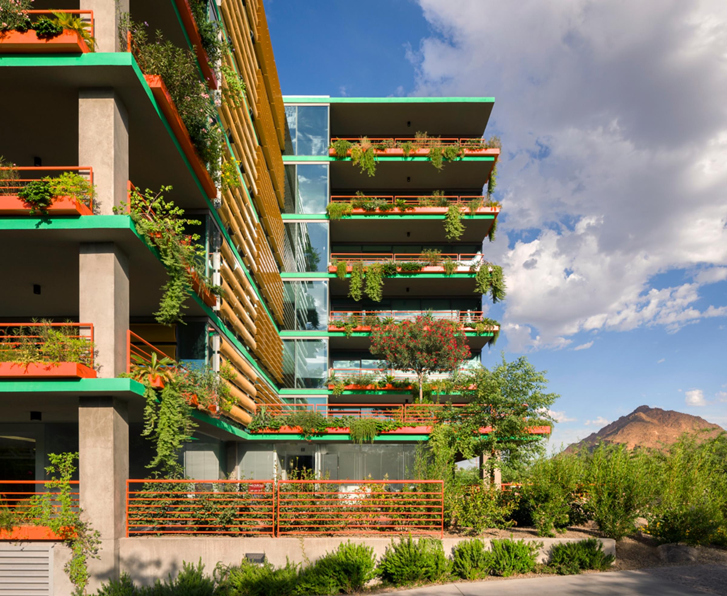 Optima Sonoran Village modern apartment building with lush green balconies and a mountainous backdrop under a partly cloudy sky.