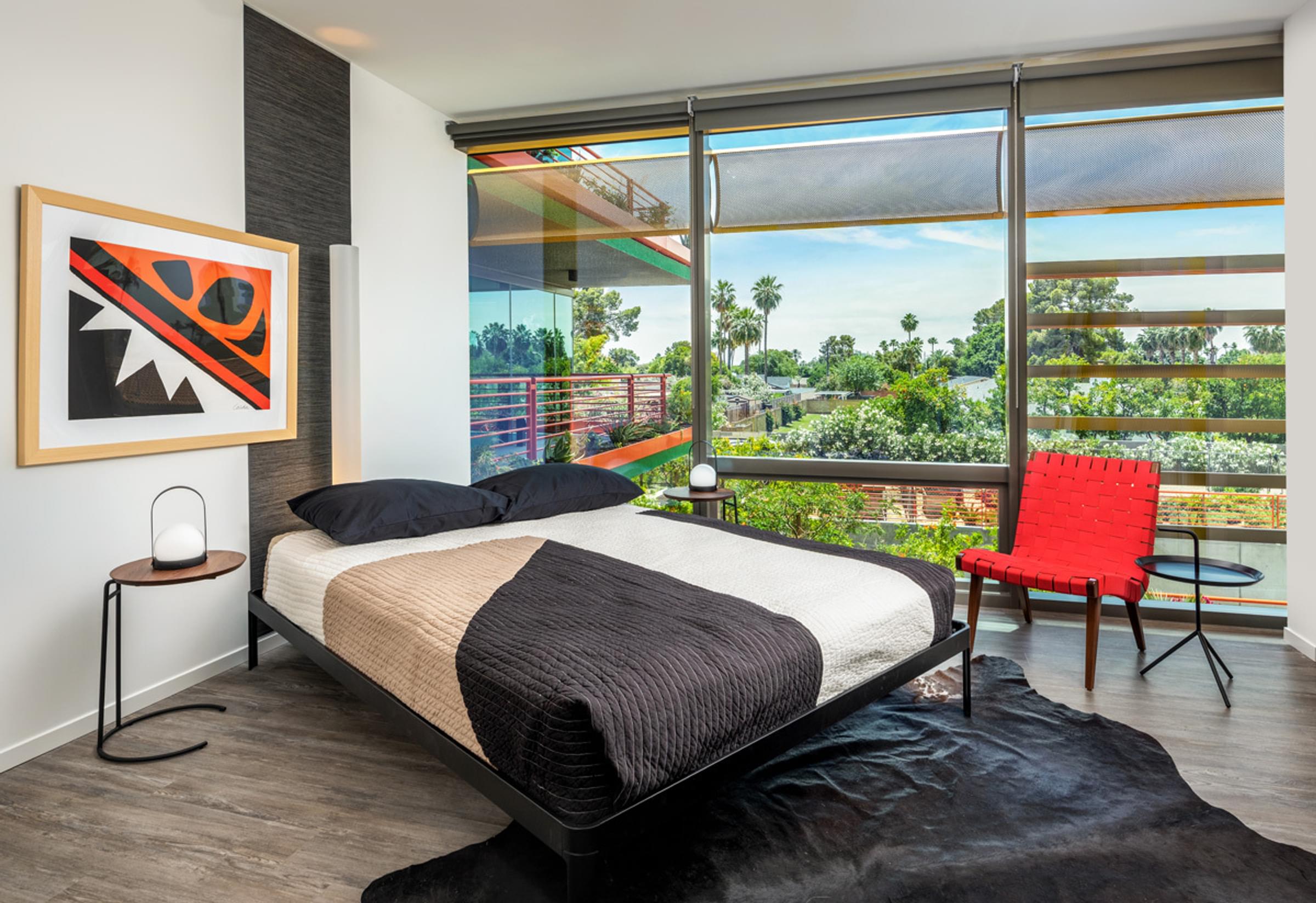 Optima Sonoran Village apartments Modern bedroom with a large window, abstract artwork, a bed with black and beige bedding, and a red chair.