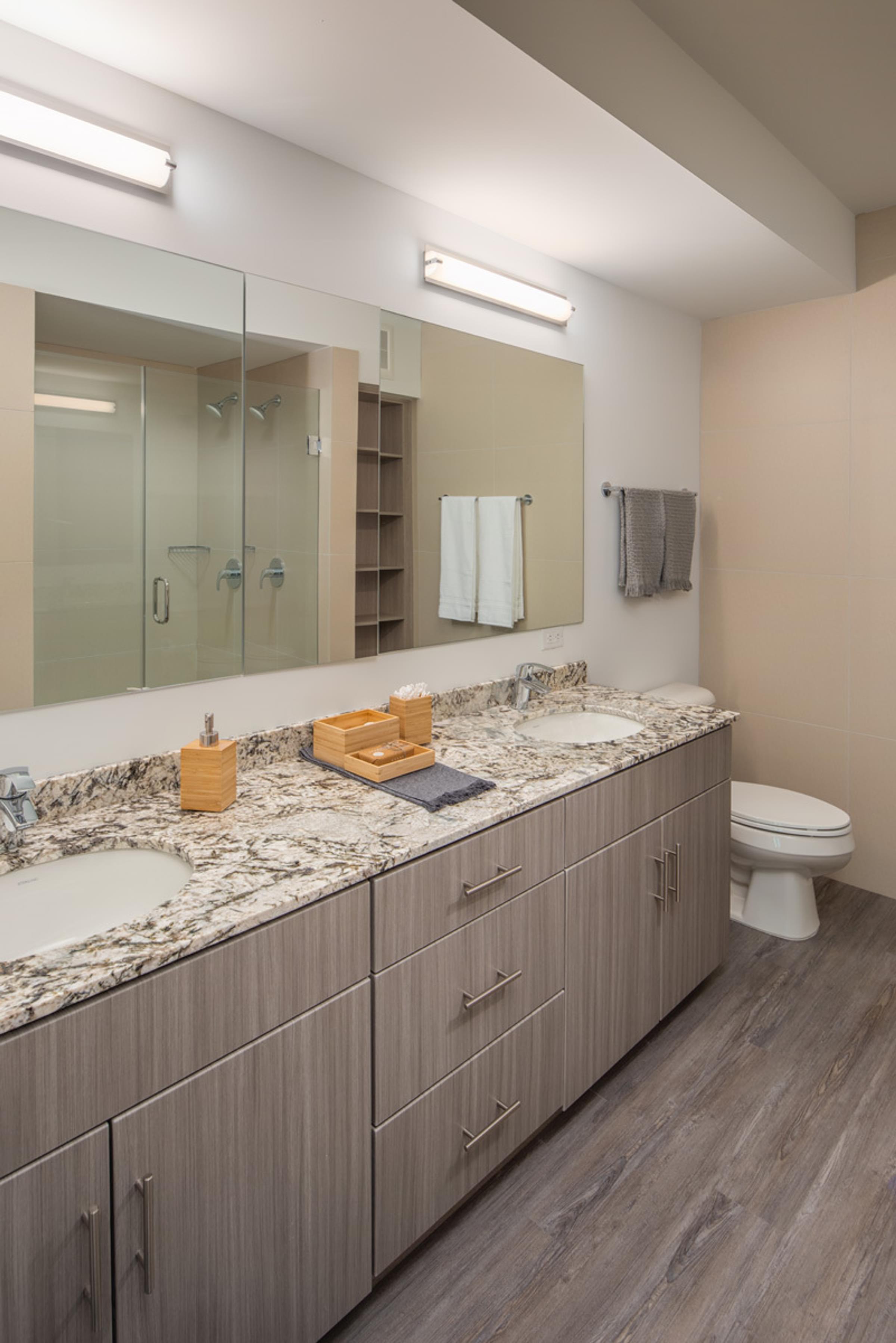 Optima Sonoran Village apartments Modern bathroom with double sink, granite countertop, large mirrors, and wood cabinets.