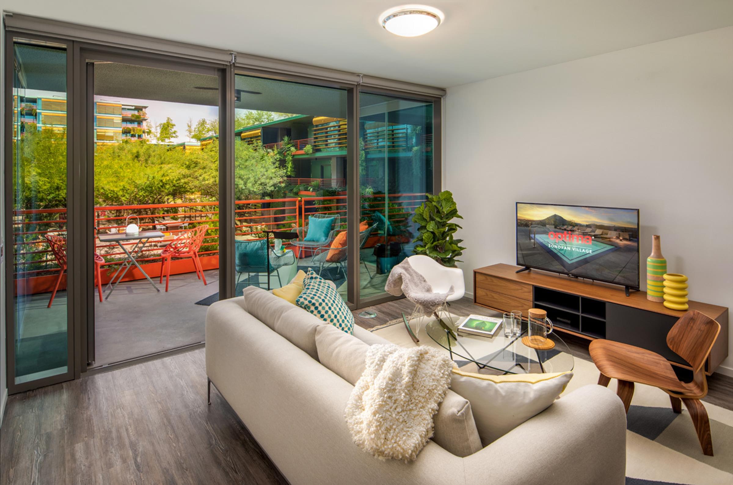 Optima Sonoran Village apartmentsModern living room with a sofa, TV, and plants. Glass doors open to a balcony with a table and chairs.