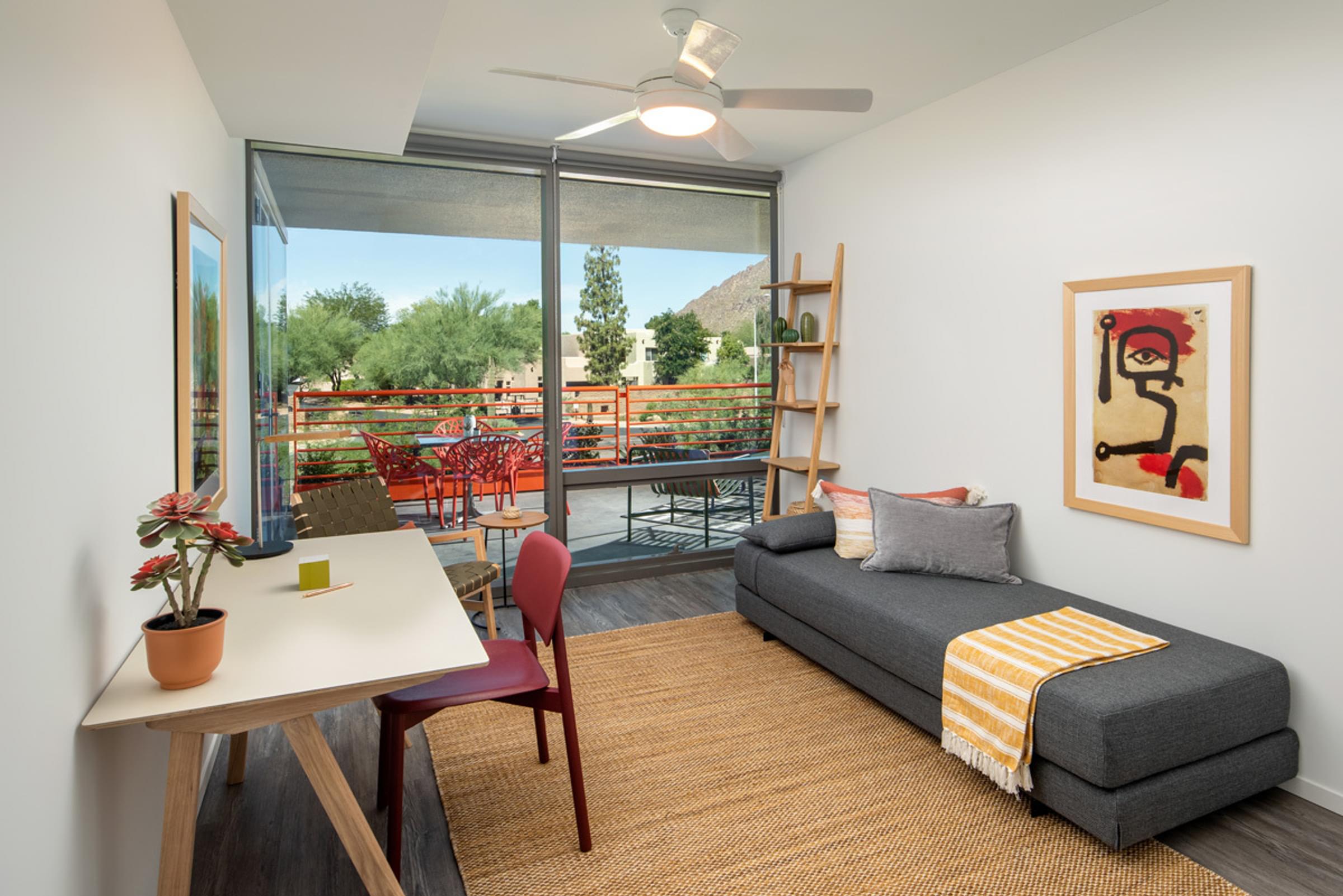 Optima Sonoran Village apartments Modern room with a daybed, desk, chair, and wall art. Glass doors open to a balcony with outdoor seating and nature views.