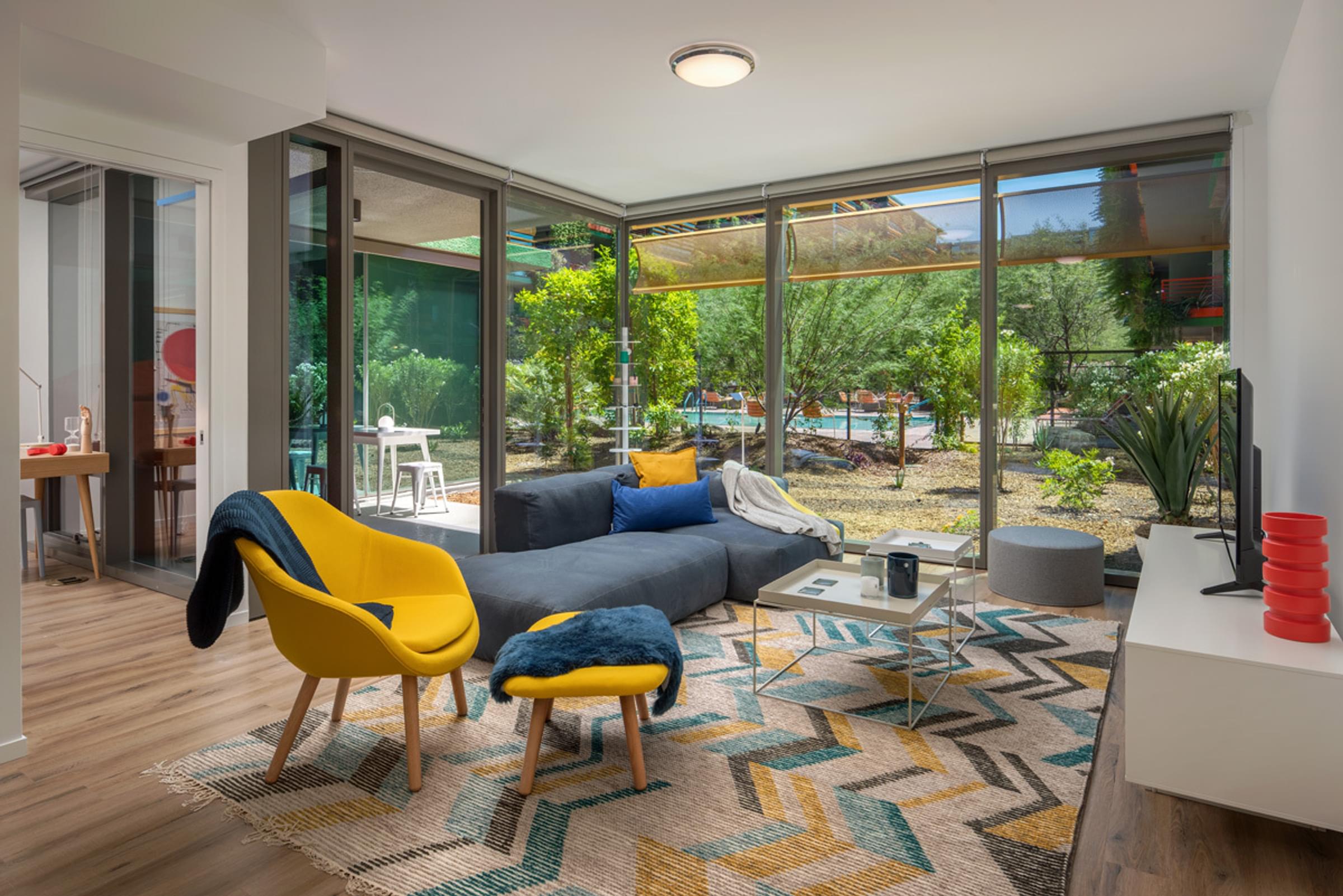 Optima Sonoran Village apartment Modern living room with large windows, yellow chairs, gray sofa, geometric rug, and view of a lush garden.