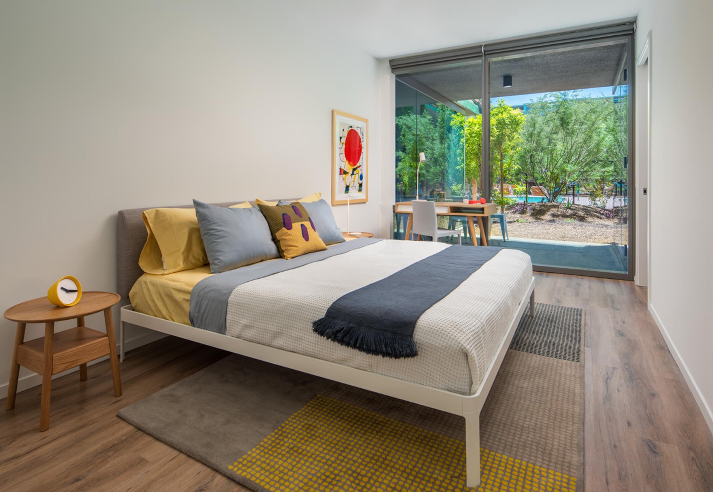 Optima Sonoran Village apartment Modern bedroom with large bed, colorful pillows, round side table, large window, and view of trees outside.