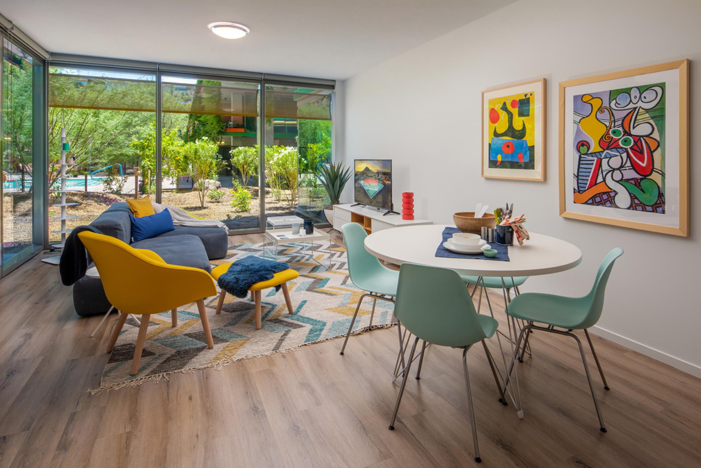 Optima Sonoran Village apartments Modern living room and dining area with colorful chairs, abstract art, large windows, and garden view.