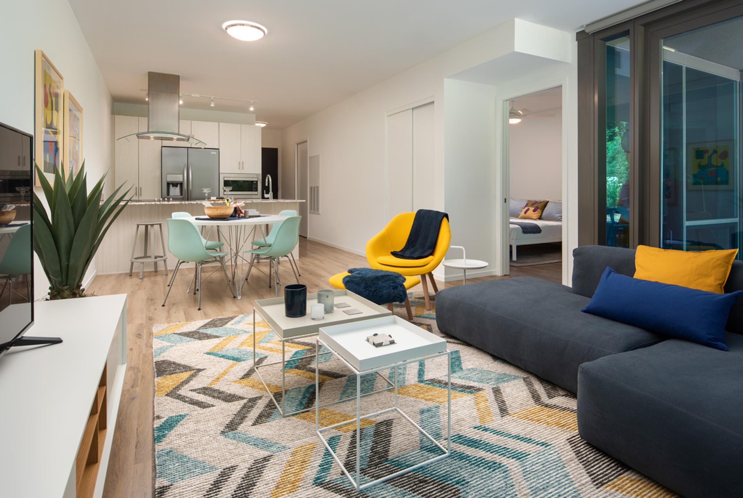 Optima Sonoran Village apartments Modern living room with a gray sofa, yellow chair, colorful rug, and a view into a kitchen and bedroom.