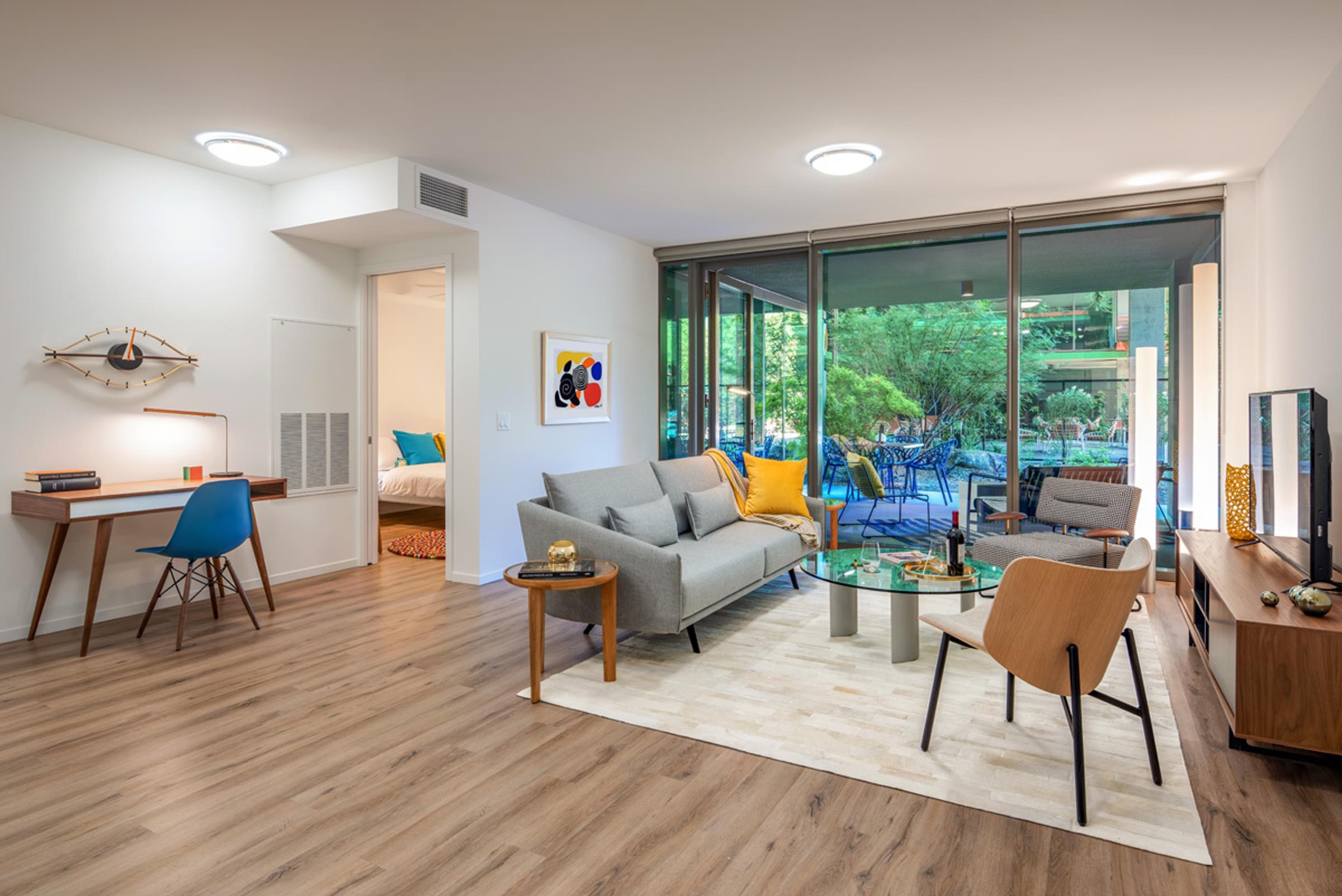 Optima Sonoran Village apartments Modern living room with large windows, gray sofa, wooden furniture, and a small desk; view of a lush garden outside.