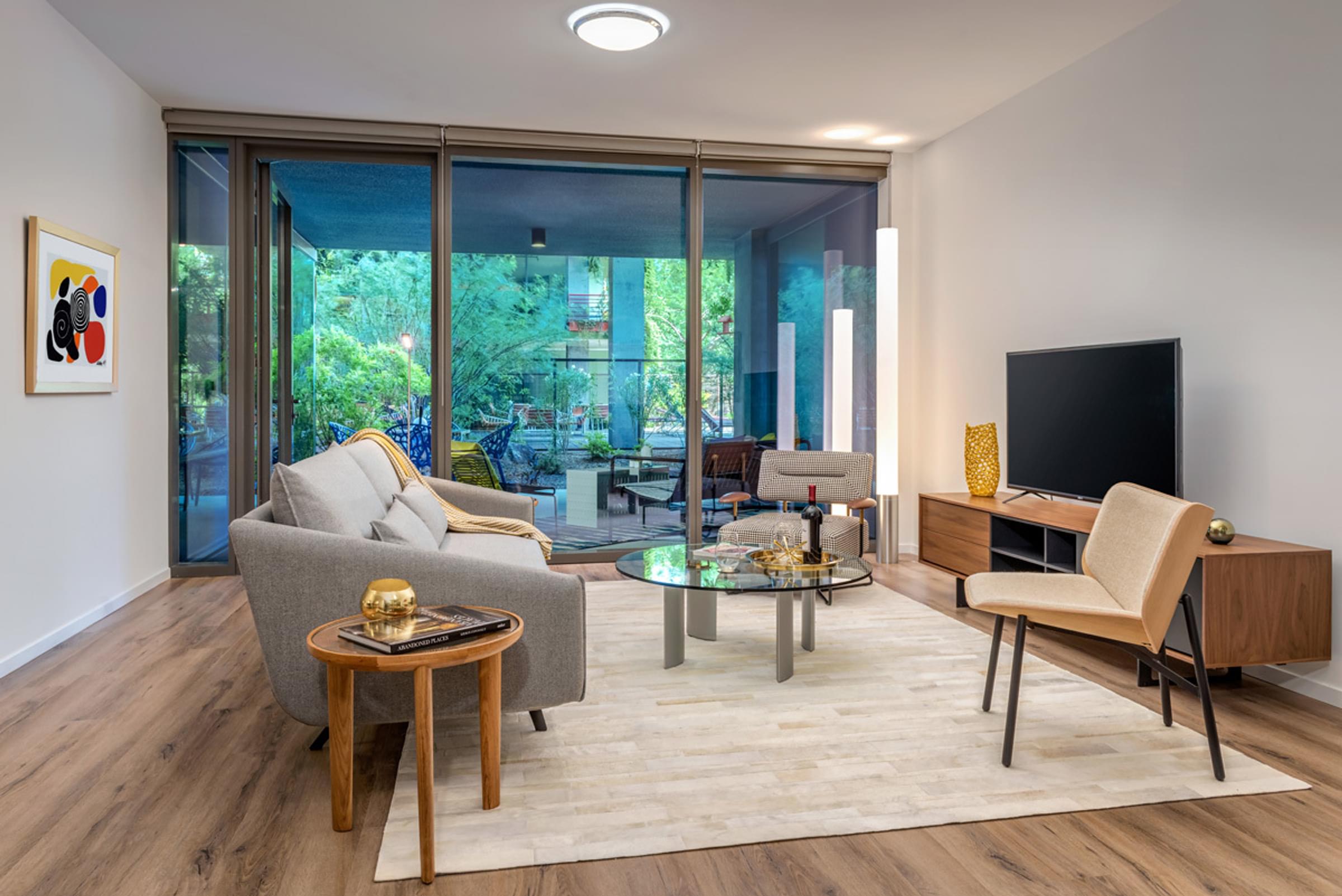 Optima Sonoran Village apartments Modern living room with glass doors, neutral sofas, a glass coffee table, and a wooden TV stand with a large screen.