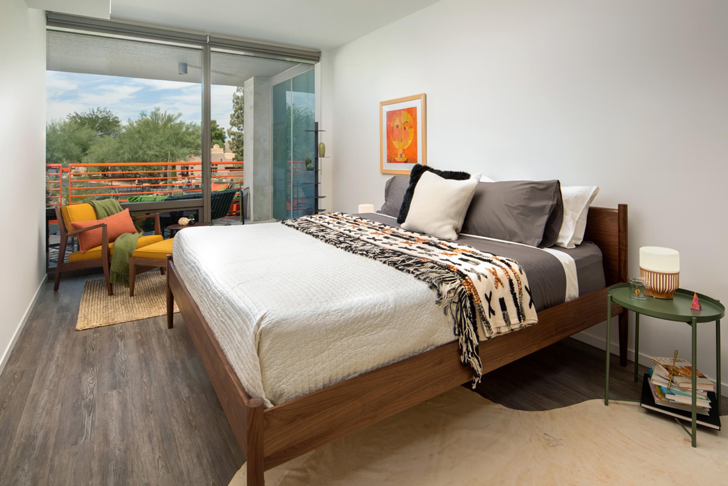 Optima Sonoran Village apartments Modern bedroom with a wooden bed, colorful pillows, and a balcony view with trees. A chair and side table are nearby.