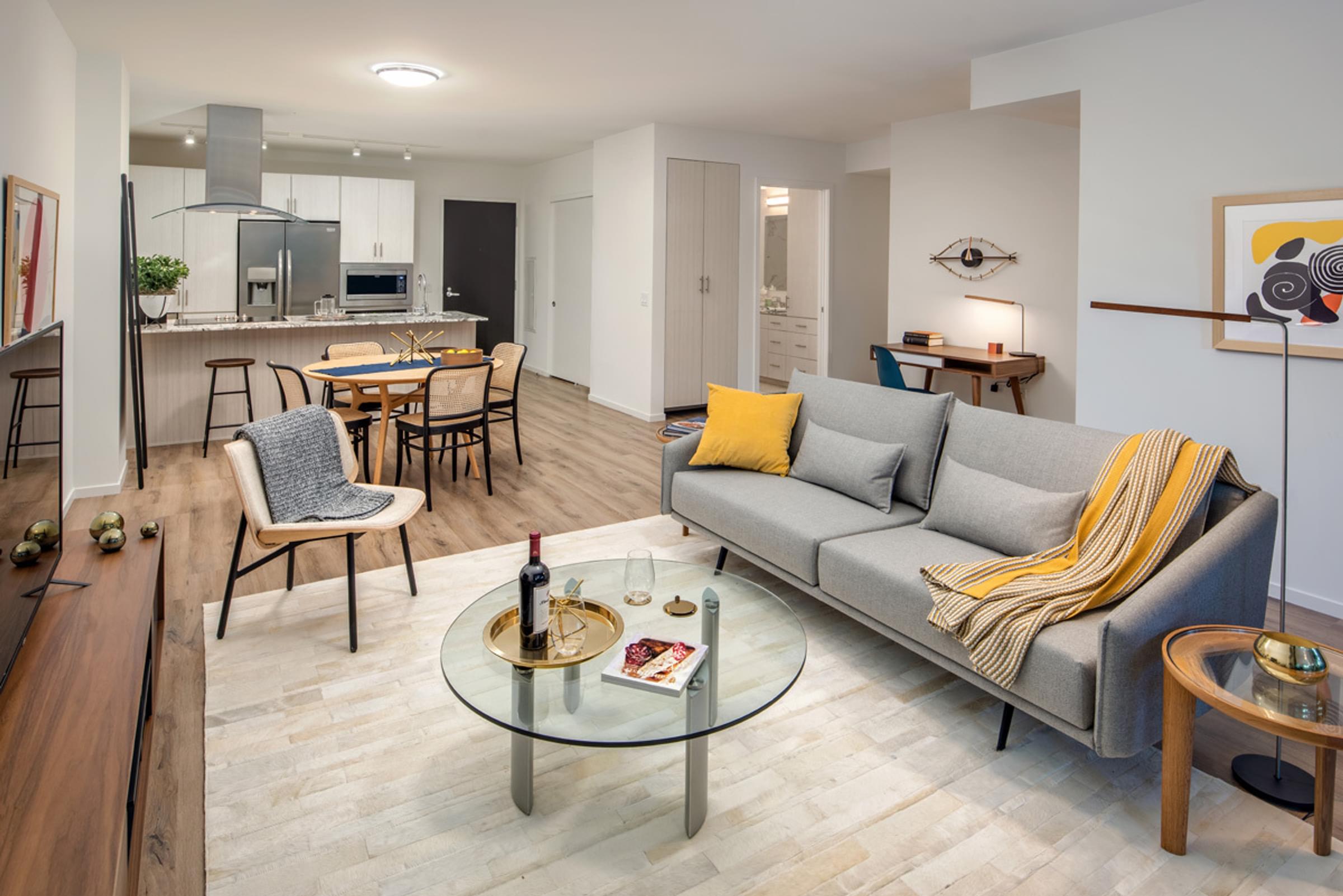 Optima Sonoran Village apartments Modern living room with a gray sofa, yellow throw, glass coffee table, and open kitchen with a dining area.
