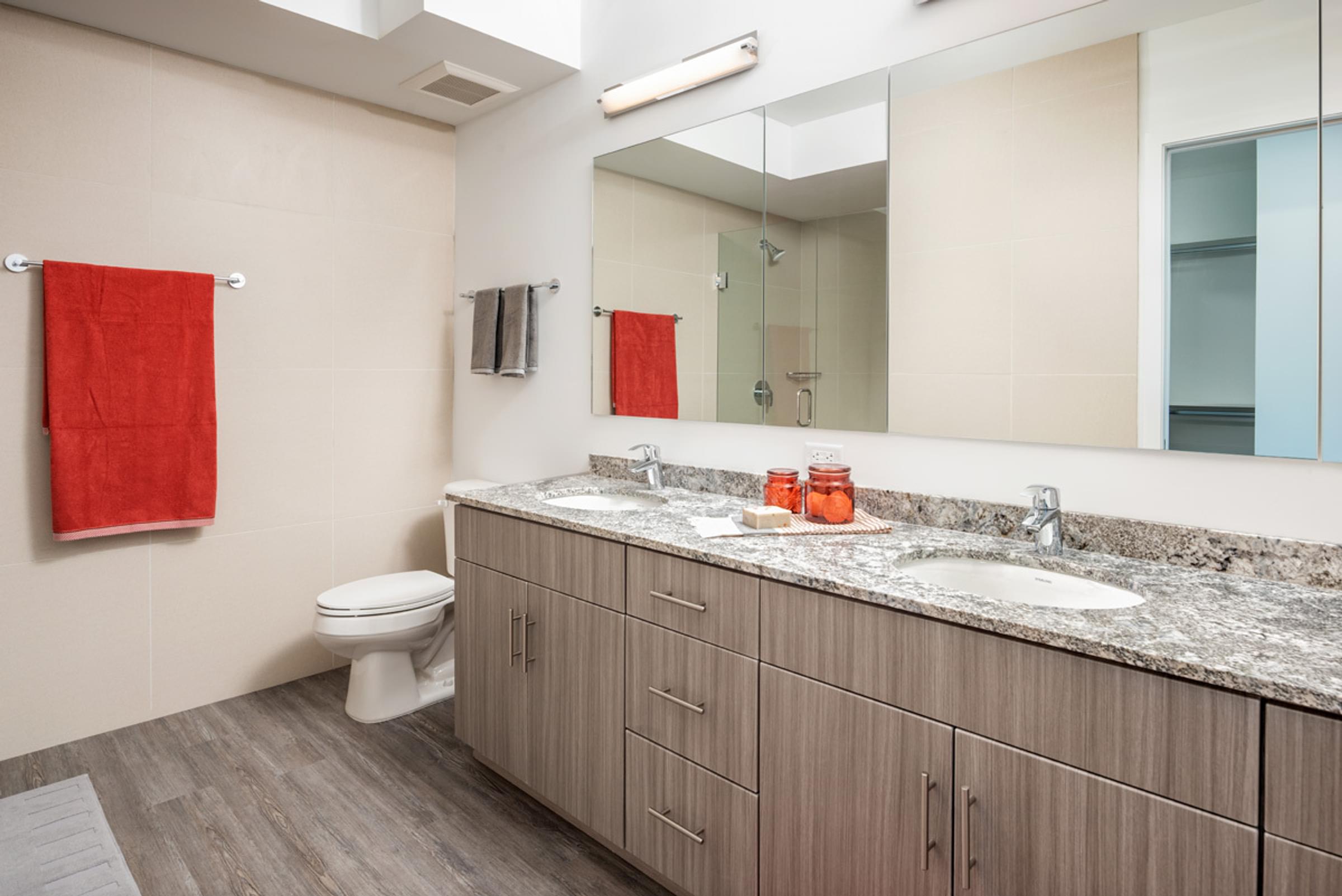 Optima Sonoran Village apartments A modern bathroom with double sinks, granite countertop, red towels, and large mirror above wooden cabinets.