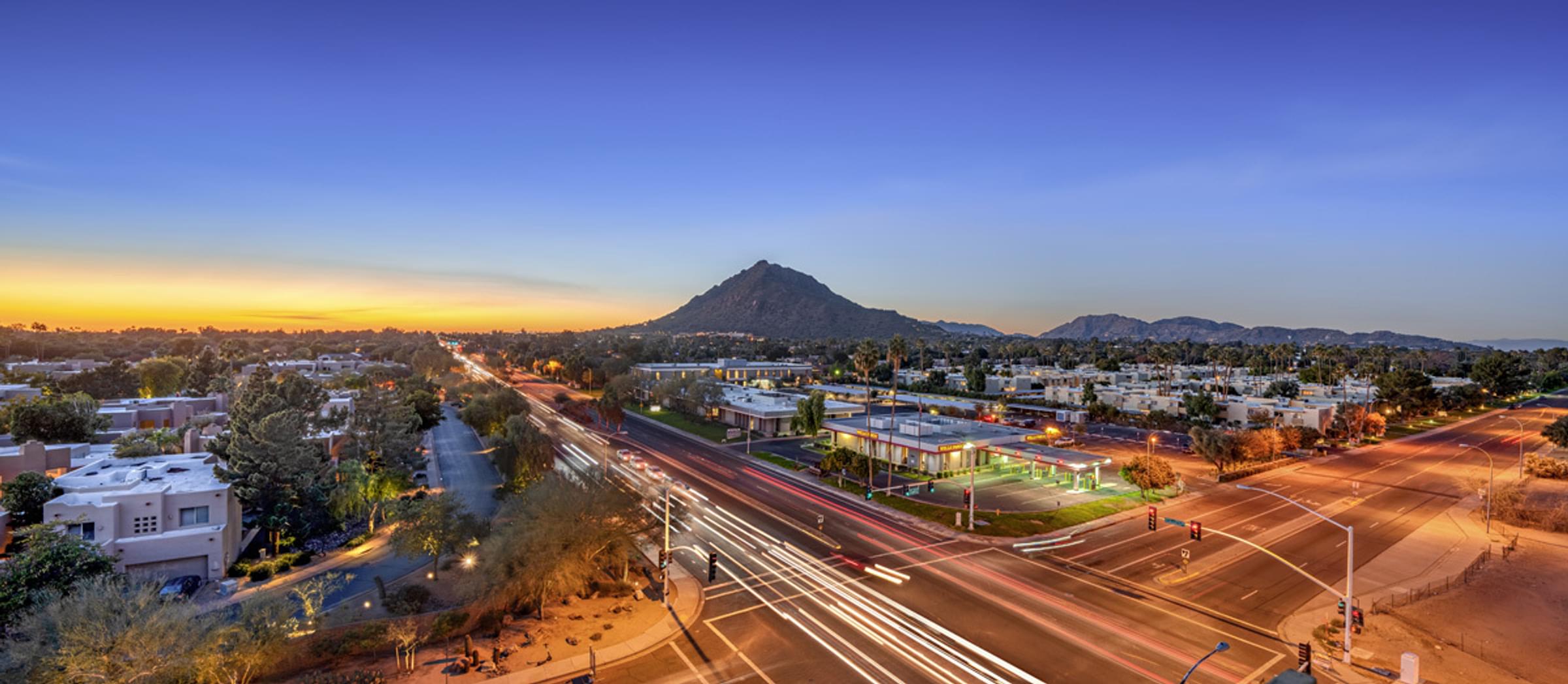 Aerial view of a city intersection at sunset, with a mountain in the background and light trails from vehicles, taken from Optima Sonoran Village apartment balcony.