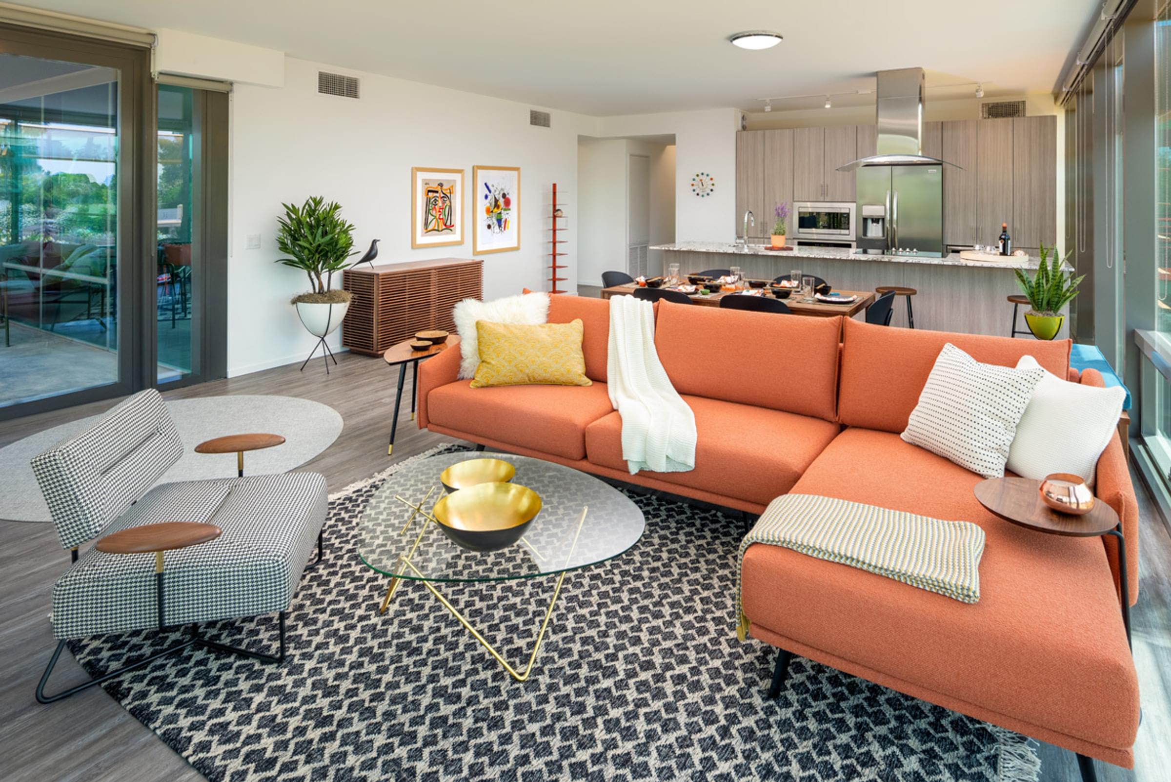 Optima Sonoran Village apartments Modern living room with an orange sectional, glass coffee table, and open kitchen. Large windows provide natural light.