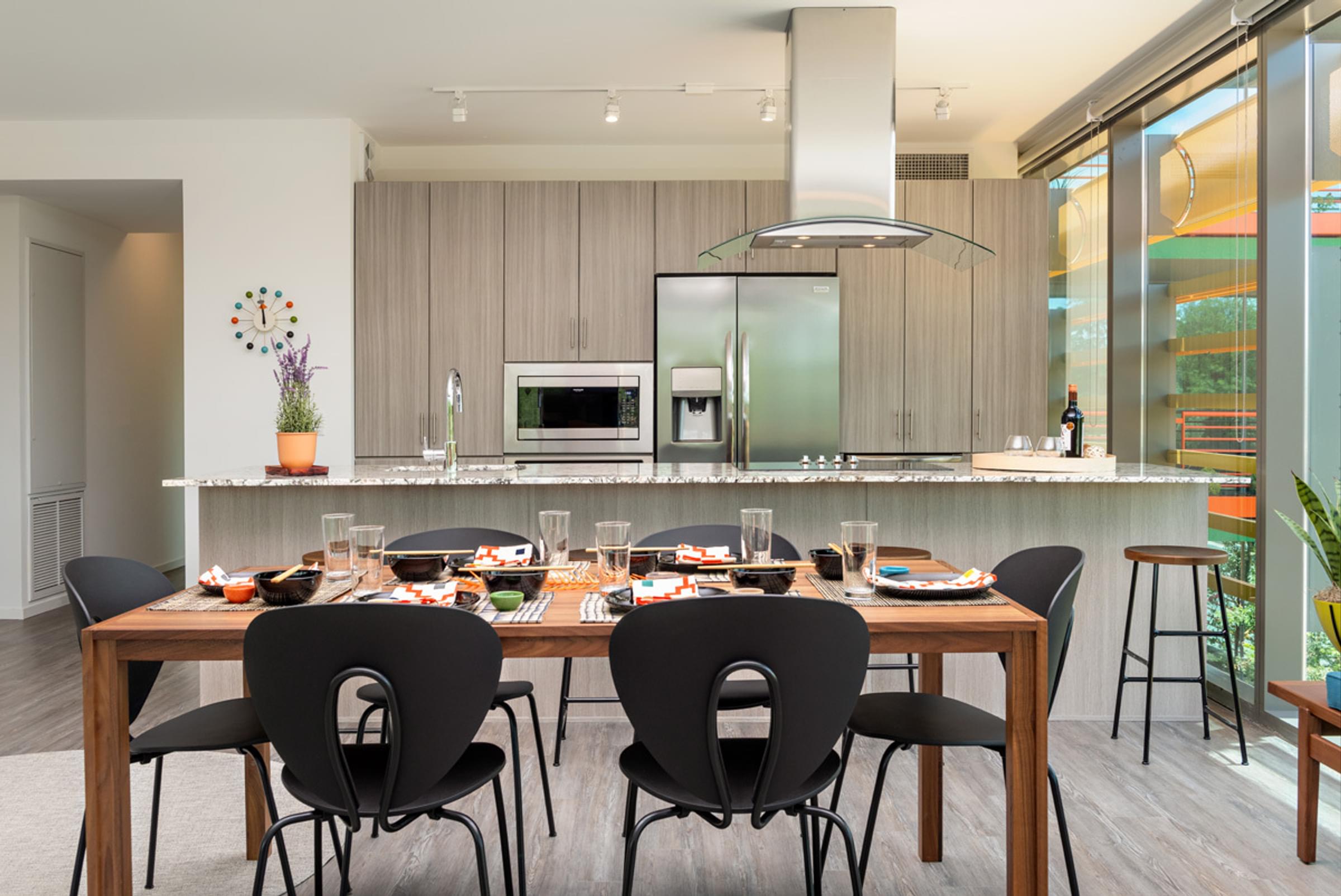 Optima Sonoran Village apartments Modern kitchen and dining area with wooden table, black chairs, and light wood cabinets. Glass doors lead outside.