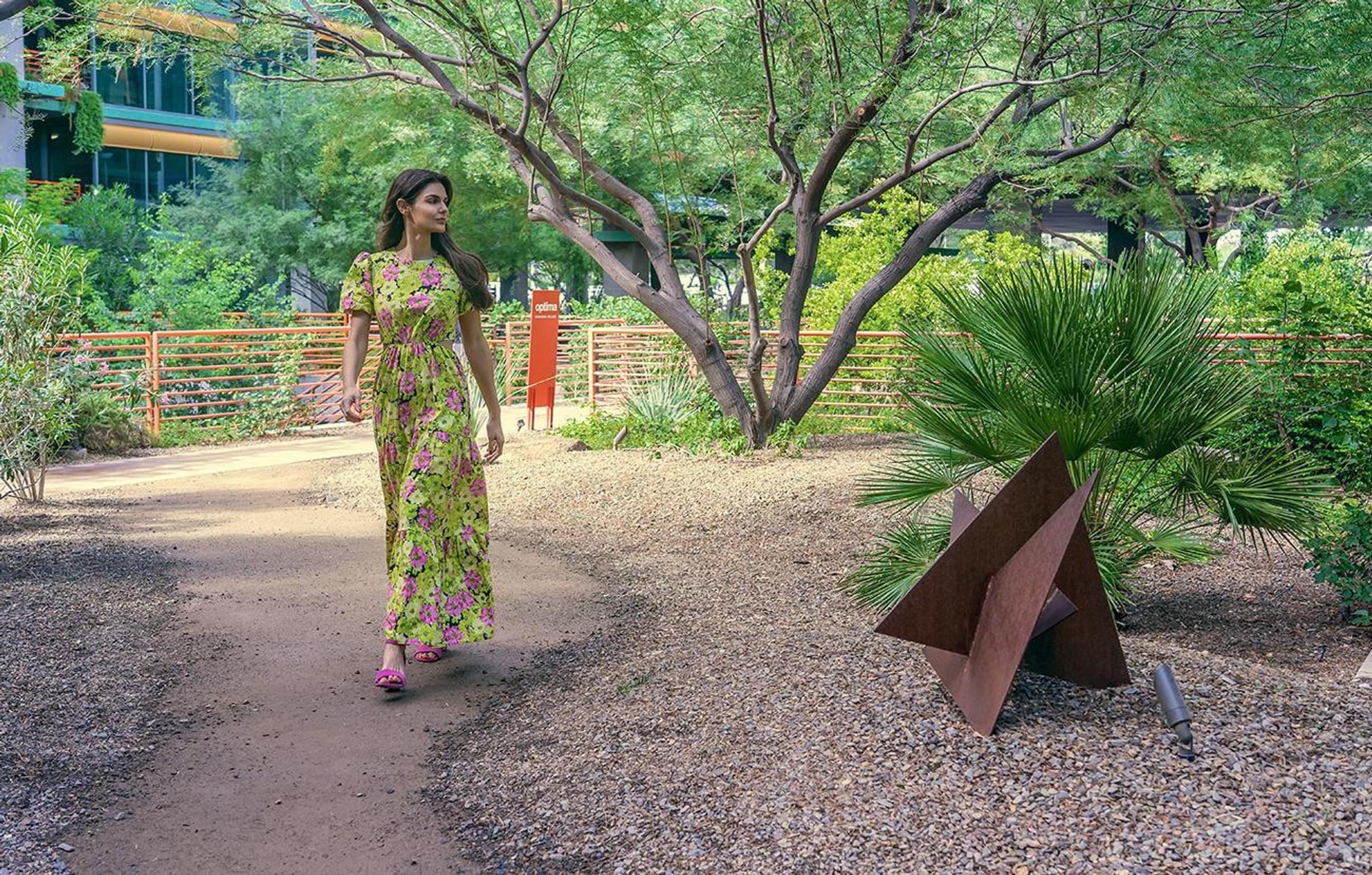 Optima Sonoran Village apartments A woman in a floral dress walks along a garden path with trees and a small metal sculpture nearby.