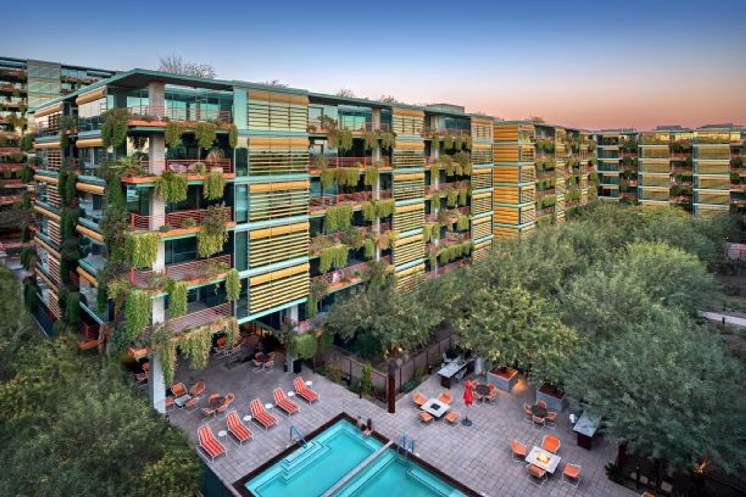 Aerial view of Optima Sonoran Village modern apartment buildings with lush balconies and a pool area surrounded by trees at sunset.