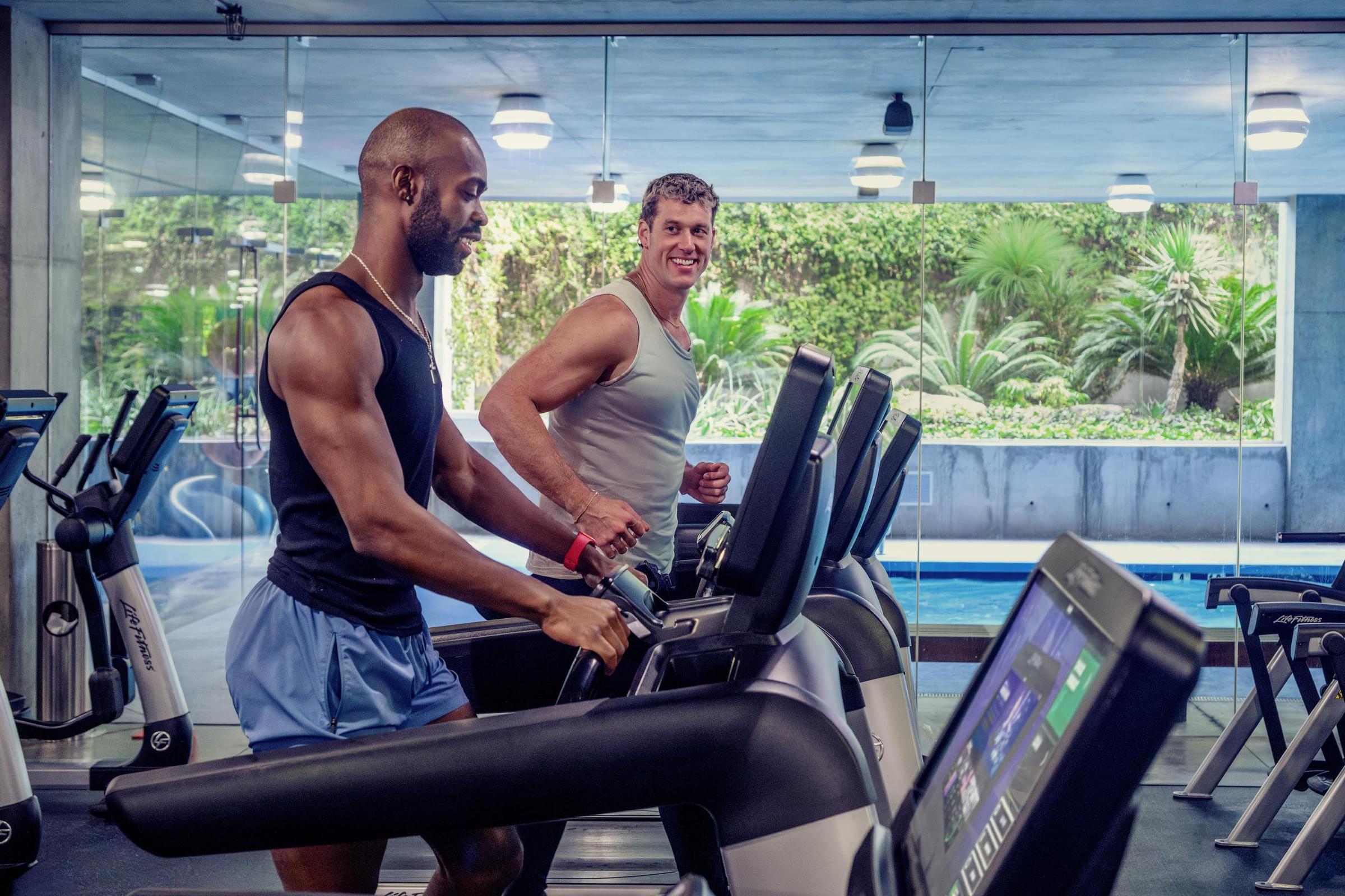 Optima Sonoran Village apartments two men exercising on treadmills in a modern gym with large windows and a view of trees outside.