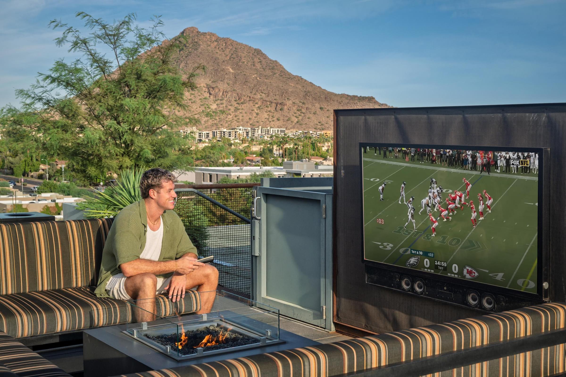 Optima Sonoran Village apartments man sitting on outdoor sofa watching a football game on a large TV with a mountain in the background.