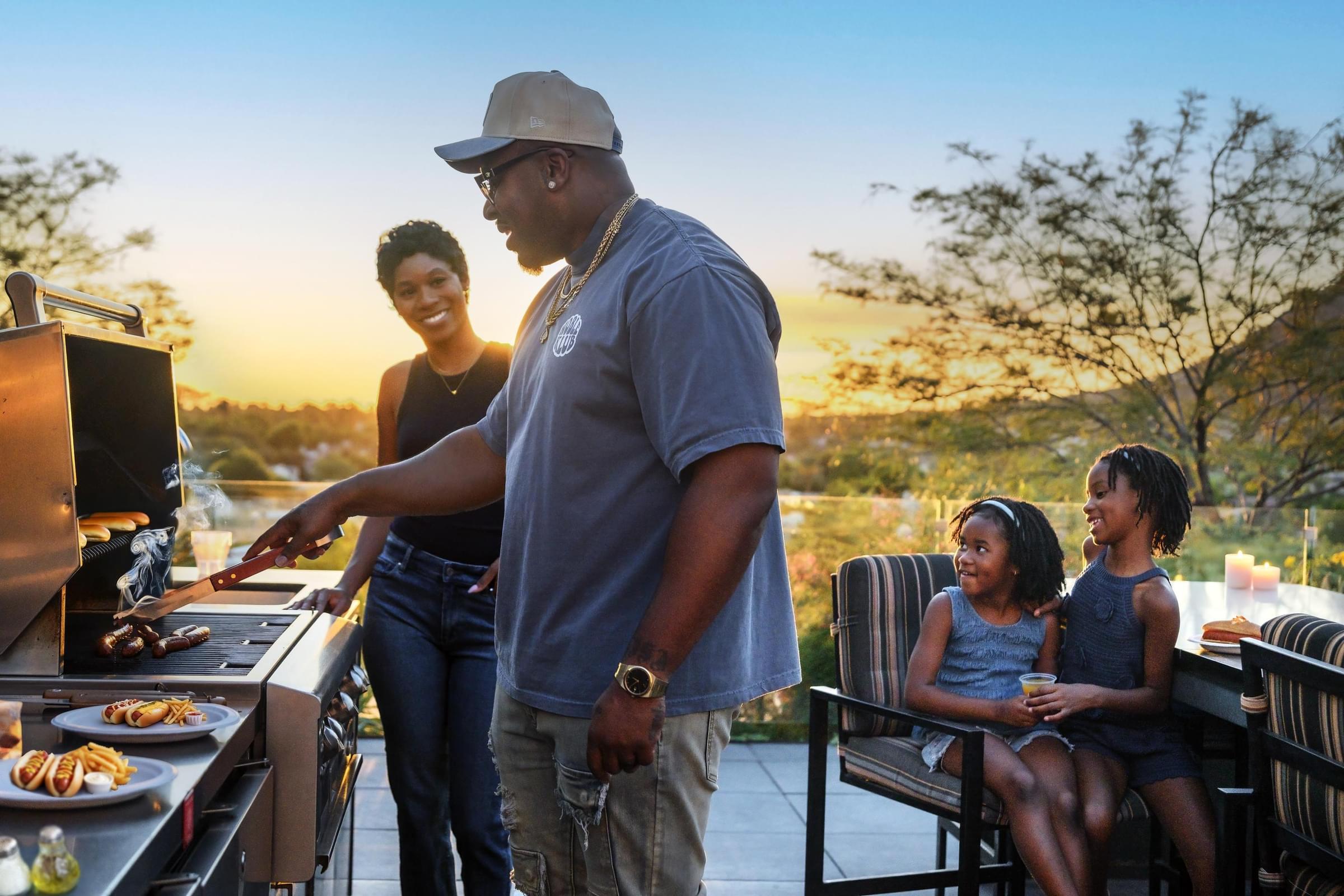 Optima Sonoran Village apartments a man grills food outdoors with a woman and two smiling children at sunset.