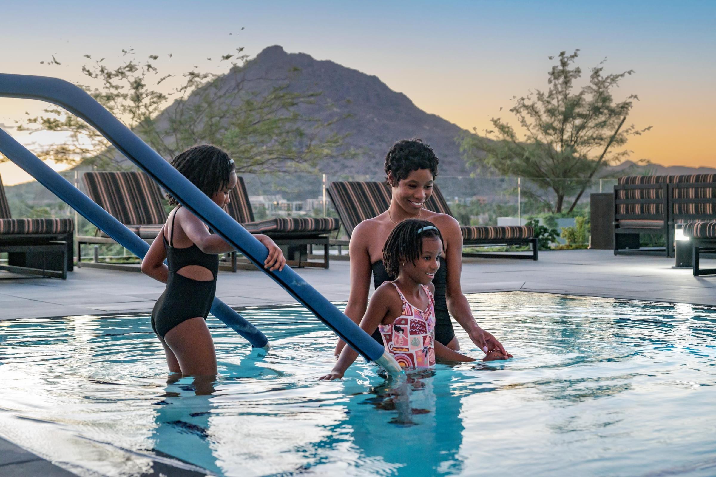 Optima Sonoran Village apartments a woman and two girls wade into a pool at sunset, with a mountain visible in the background.