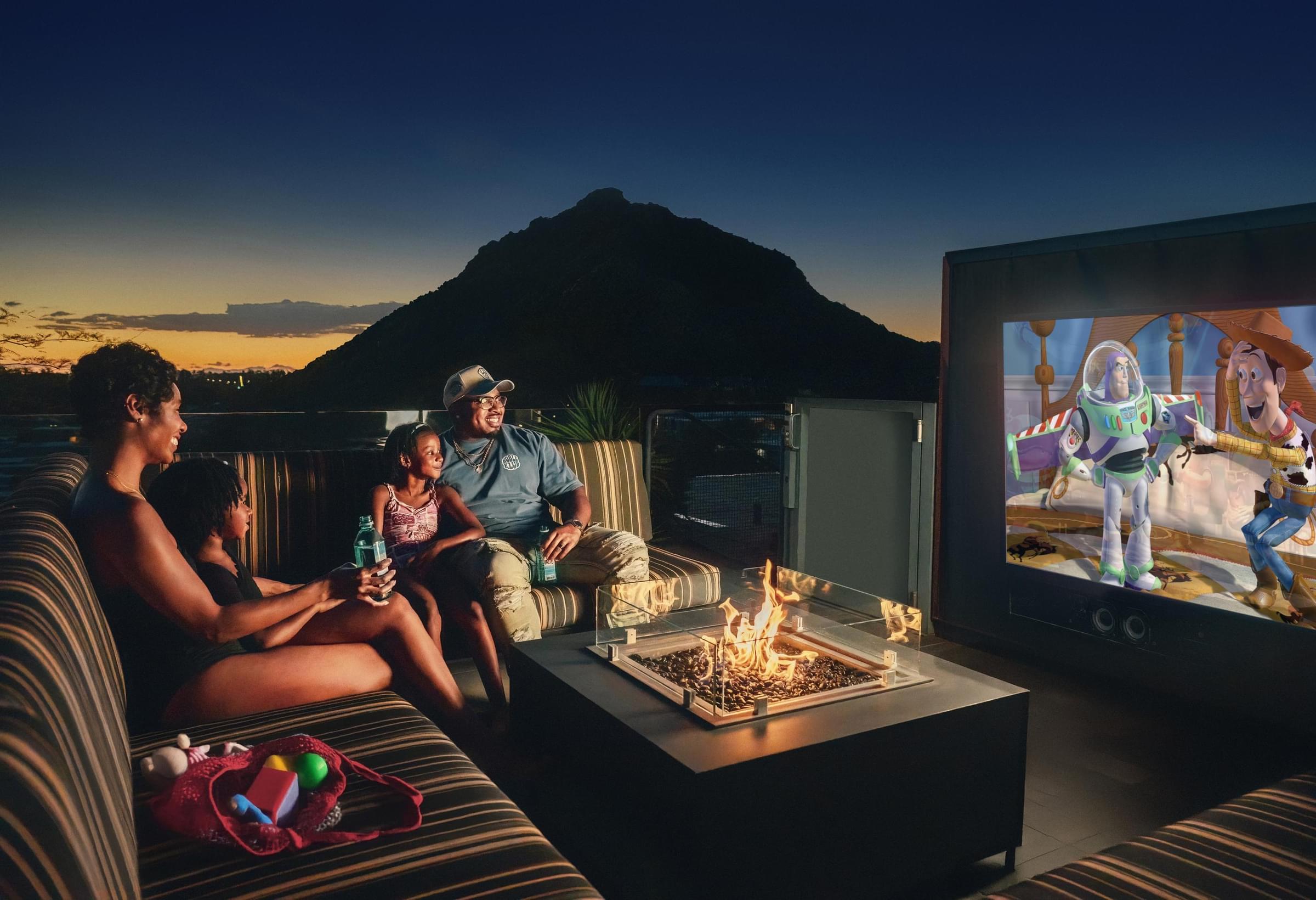 Optima Sonoran Village apartments family sits by a firepit outdoors at sunset, watching Toy Story on a large screen with a mountain behind them.