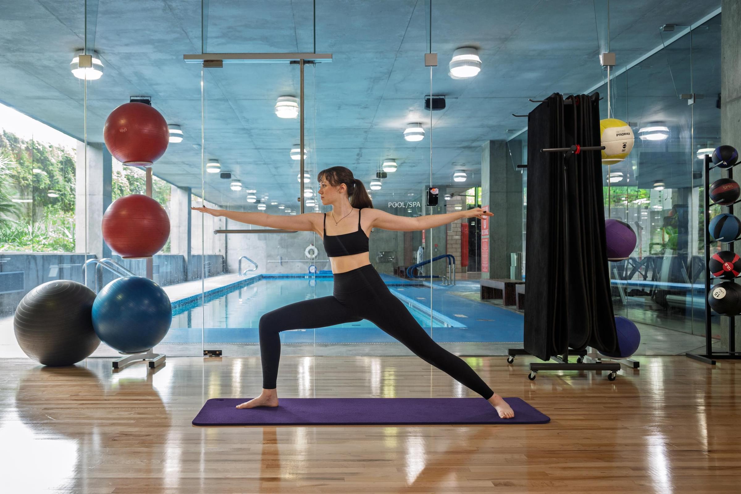 Optima Sonoran Village apartments woman in workout clothes doing yoga on a mat by an indoor pool with gym equipment nearby.