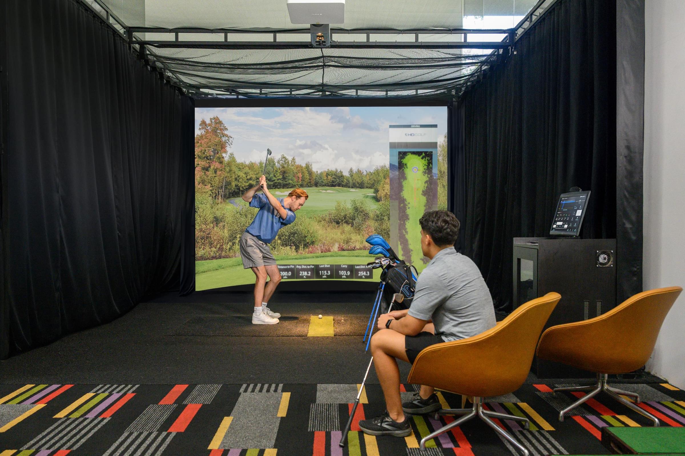 Optima Sonoran Village apartments two people use an indoor golf simulator; one swings a club while the other watches from a chair.