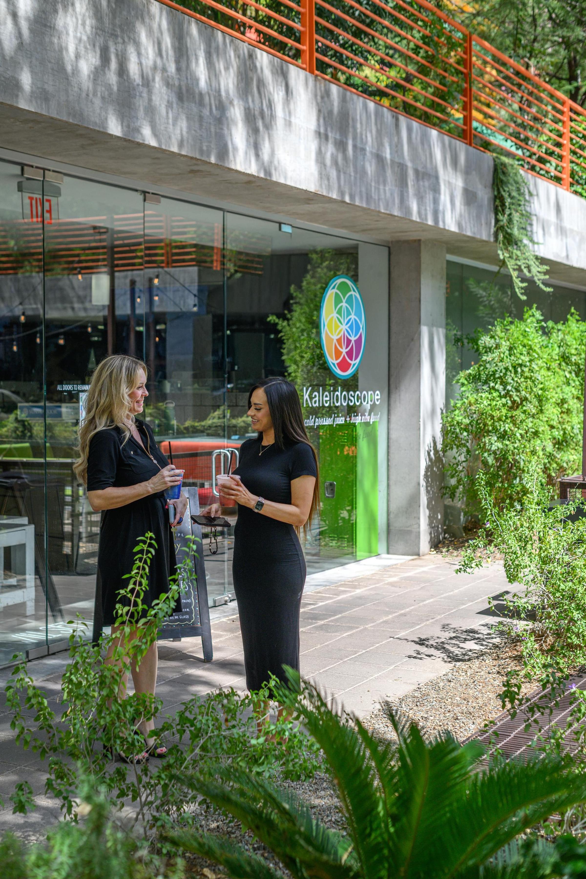 Optima Sonoran Village apartments two women standing outside Kaleidoscope juice bar, talking and holding drinks, surrounded by greenery.