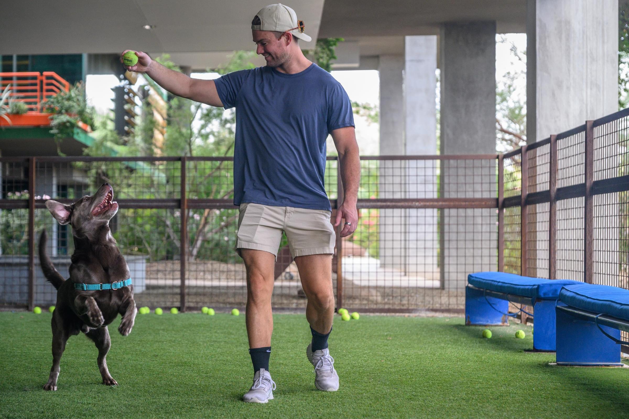 Optima Sonoran Village apartments man in a cap plays fetch with a happy dog, holding a tennis ball on a grassy, fenced area.