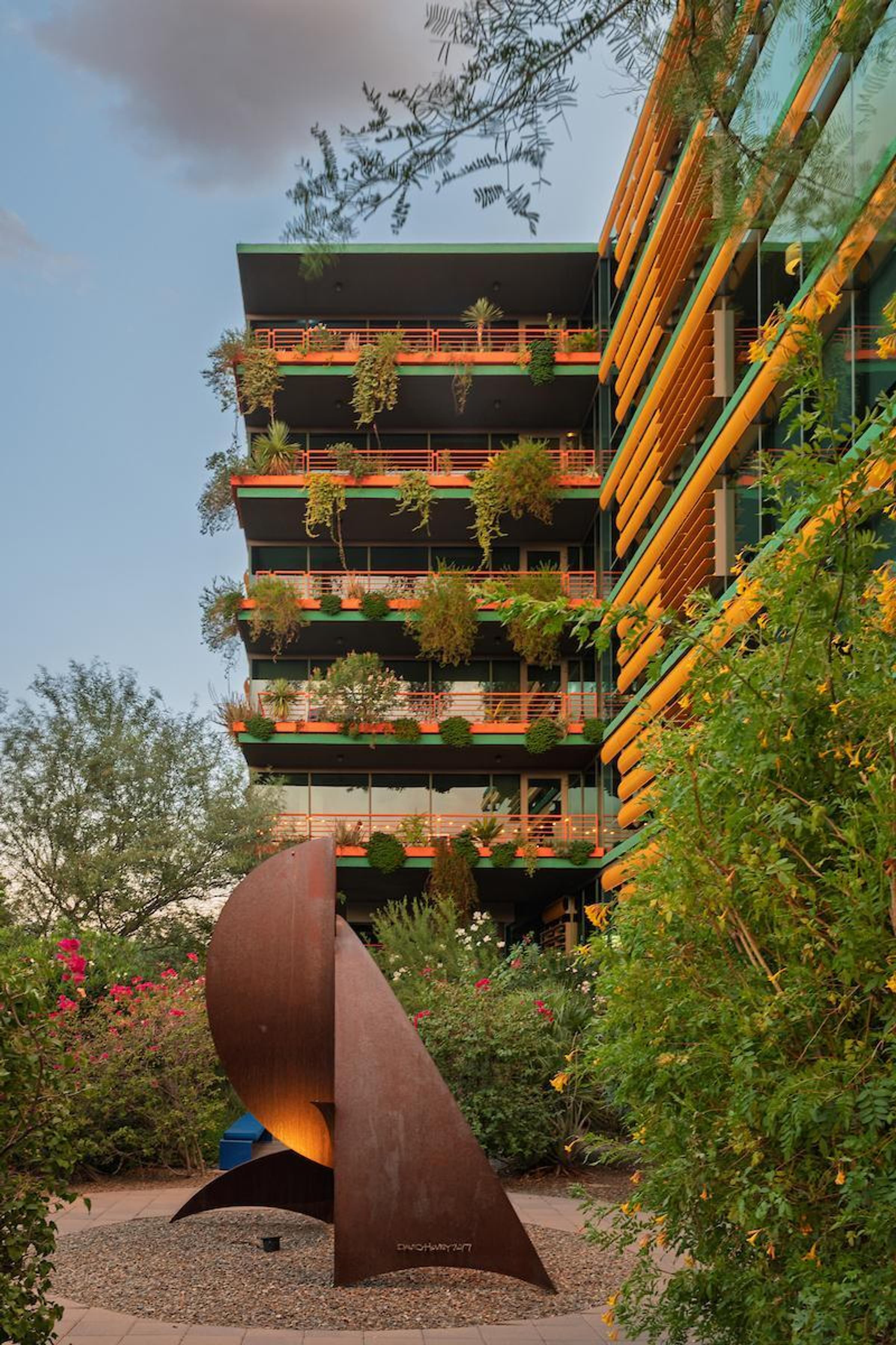Optima Sonoran Village apartments green modern building with plants on balconies and a curved metal sculpture in the landscaped foreground.