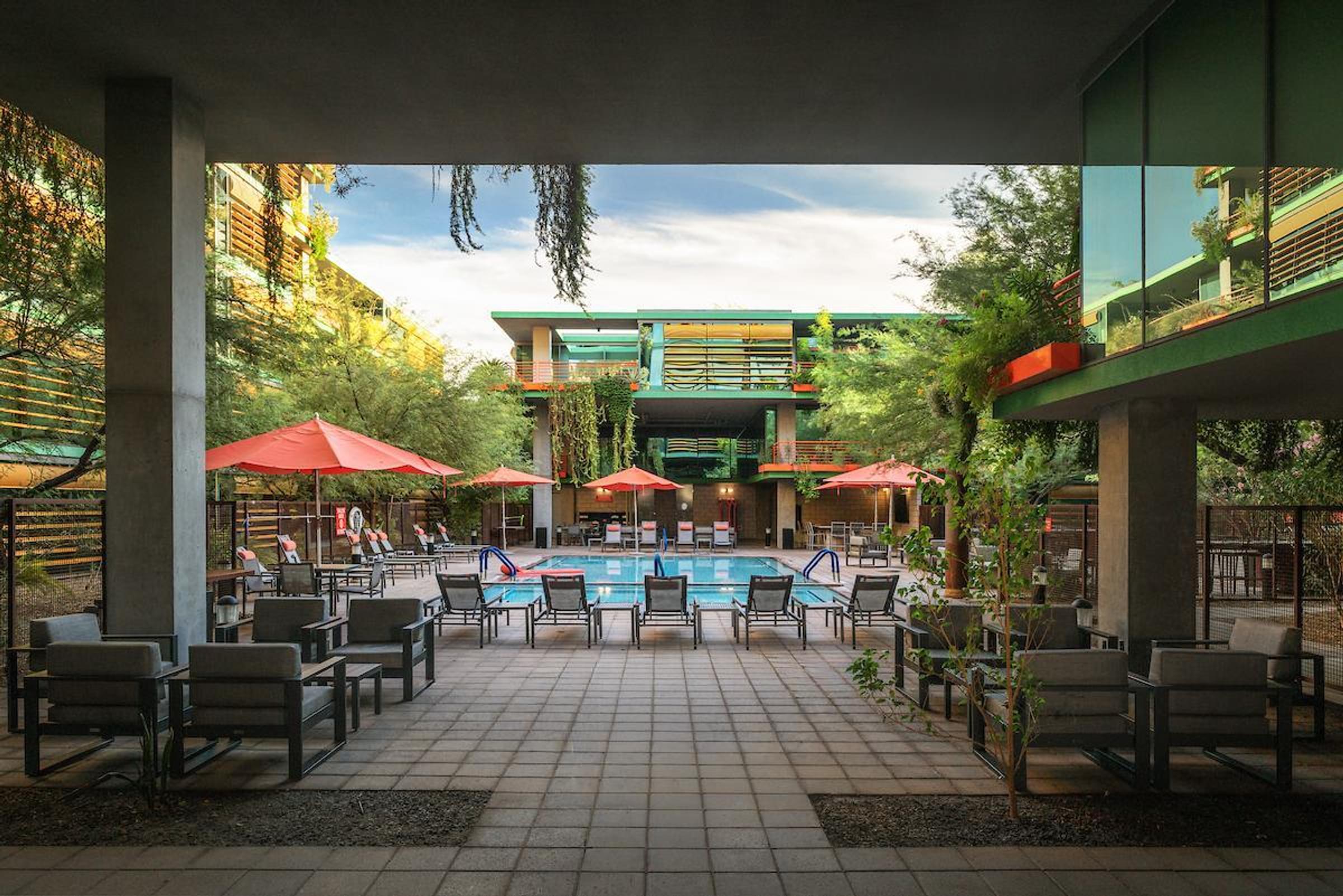 Optima Sonoran Village apartments outdoor pool area with lounge chairs, tables, and red umbrellas, surrounded by modern buildings and greenery.