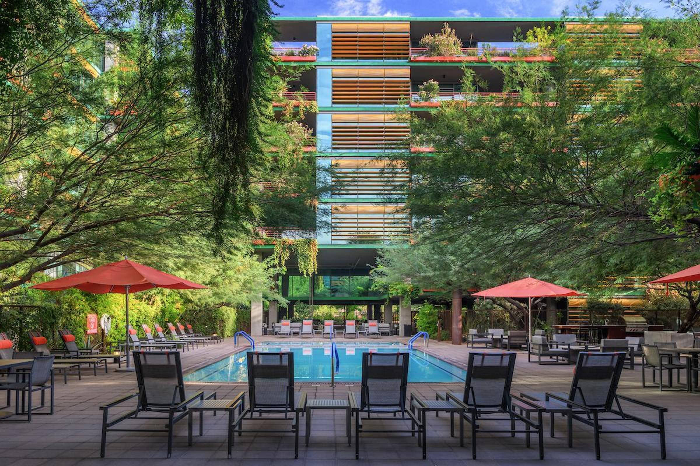 Optima Sonoran Village apartments modern apartment building with balconies, pool, lounge chairs, and red umbrellas surrounded by lush greenery.